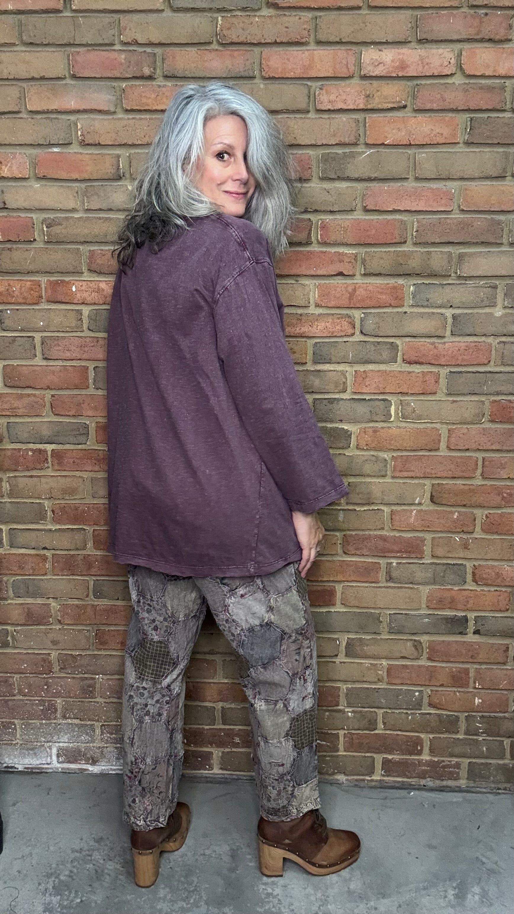 Person wearing a purple jacket and patterned pants standing against a brick wall.