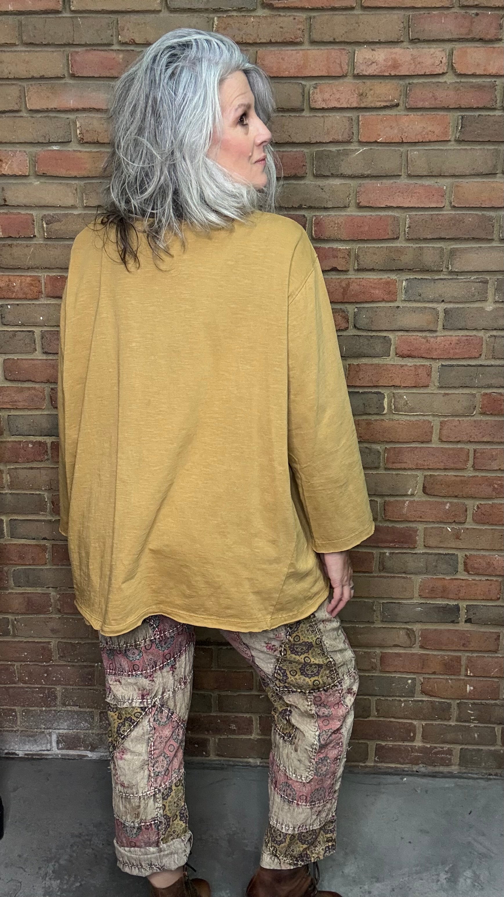 Person wearing a mustard yellow top and patterned pants standing against a brick wall.