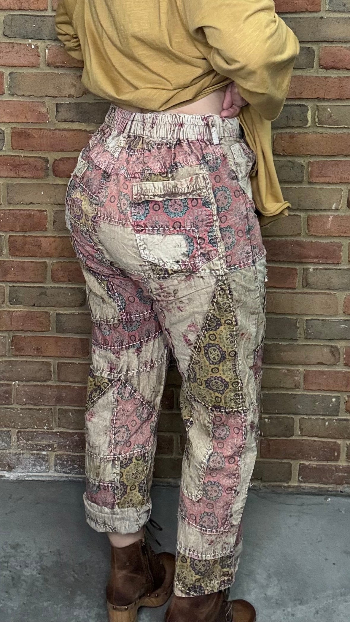 Person wearing a yellow jacket and patterned pants leaning against a brick wall.