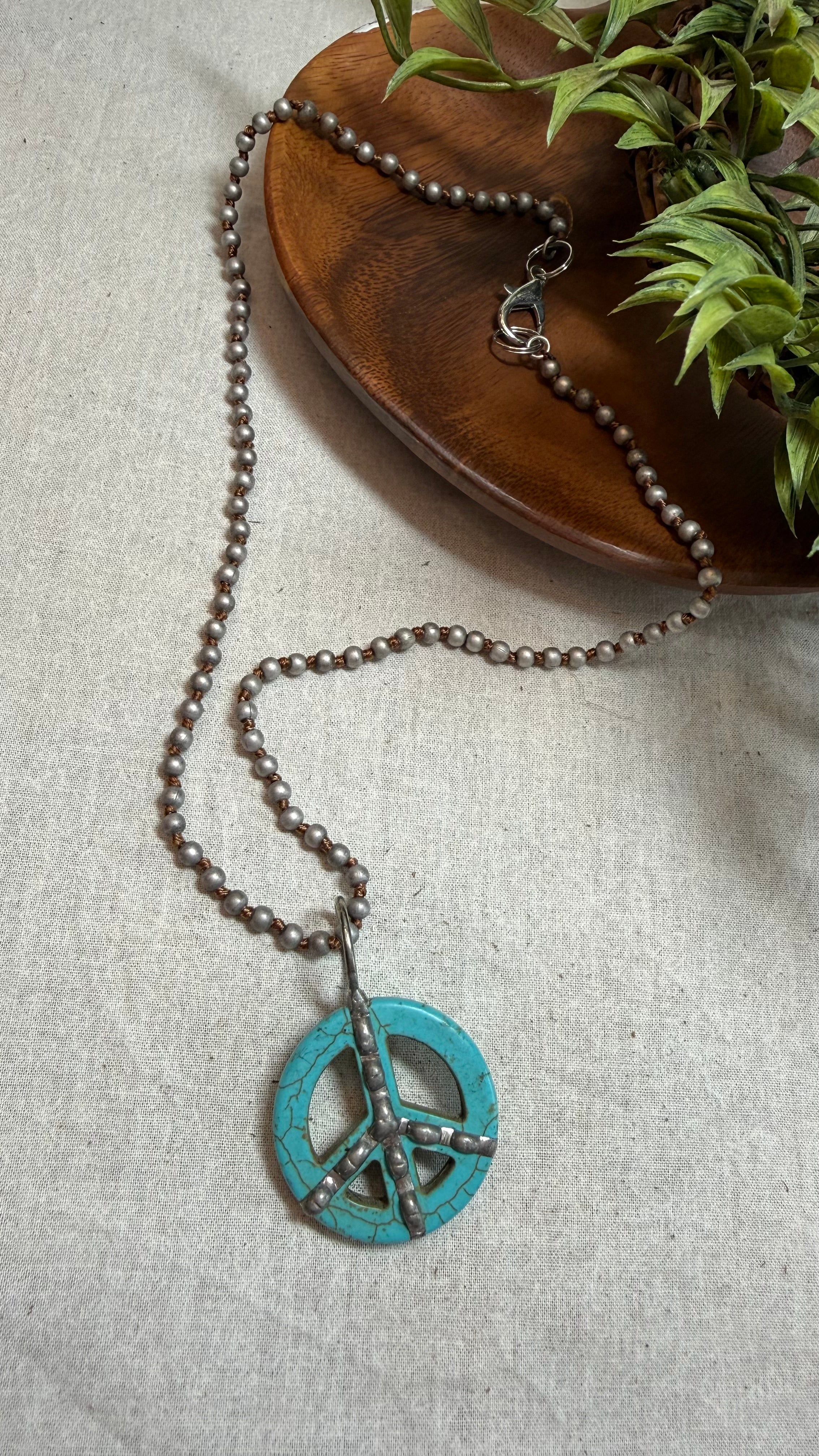 A beaded necklace with a turquoise peace sign pendant, styled on a light beige linen fabric next to a wooden dish and green leaves.