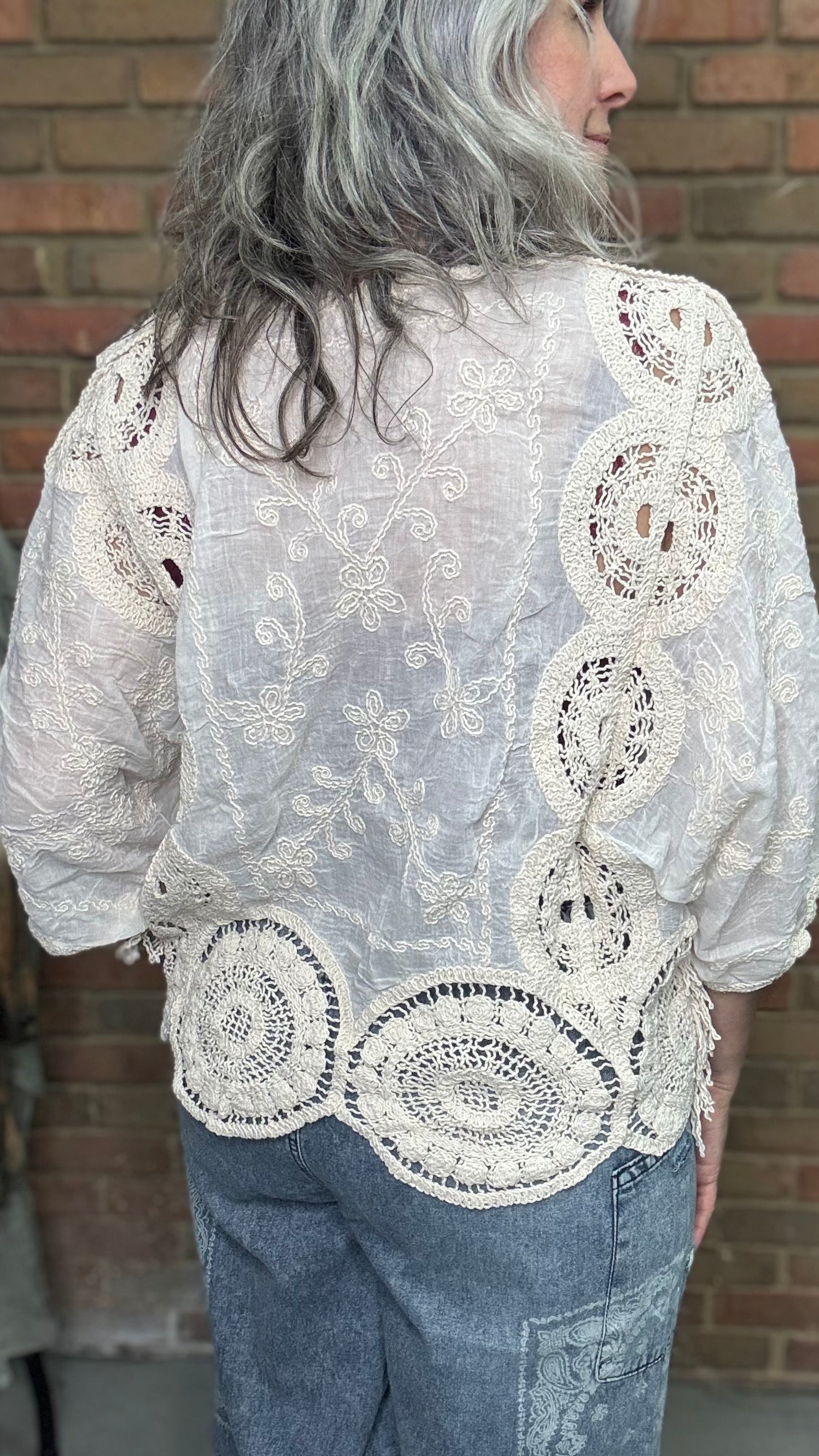 Person wearing a white lace top and blue jeans against a brick wall.