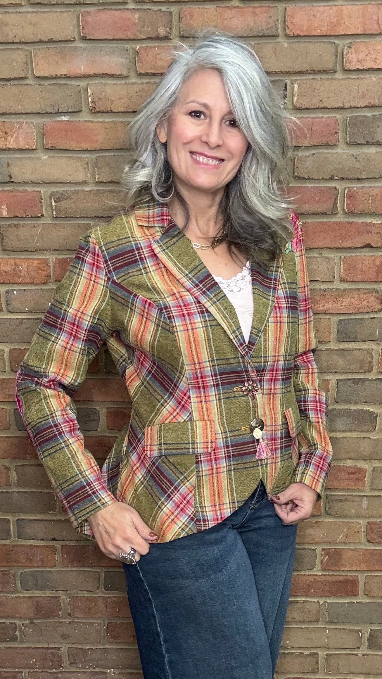 Woman wearing a plaid jacket and jeans standing against a brick wall.