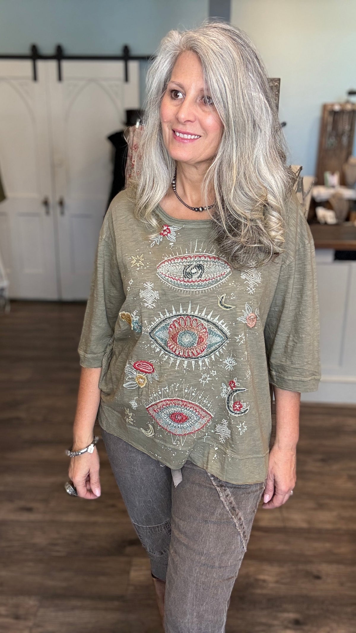 A woman with long gray hair wearing a sage green bohemian-style t-shirt with colorful embroidered eye and floral patterns, paired with gray jeans, standing in a boutique setting.