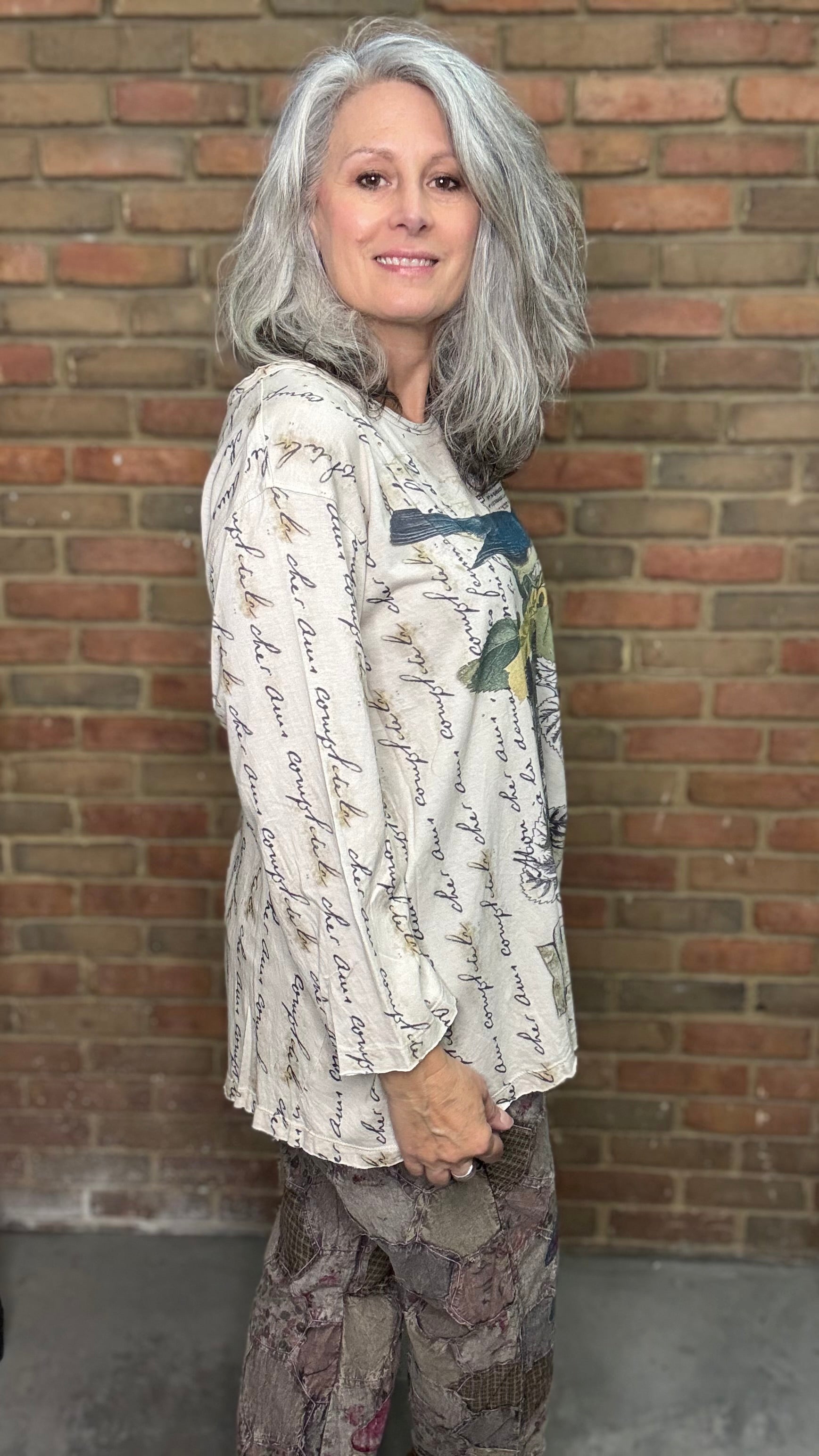 Woman wearing a patterned blouse and pants standing against a brick wall.