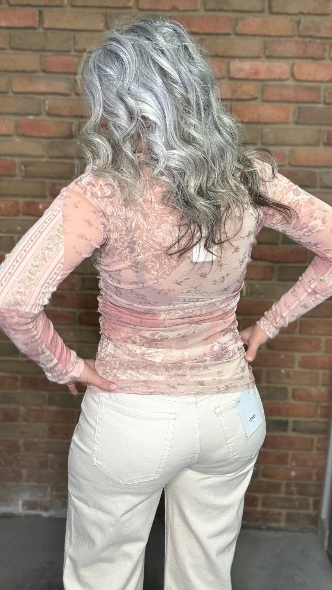 Woman wearing Mix Print Long Sleeve Mesh Top and white pants standing against a brick wall.
