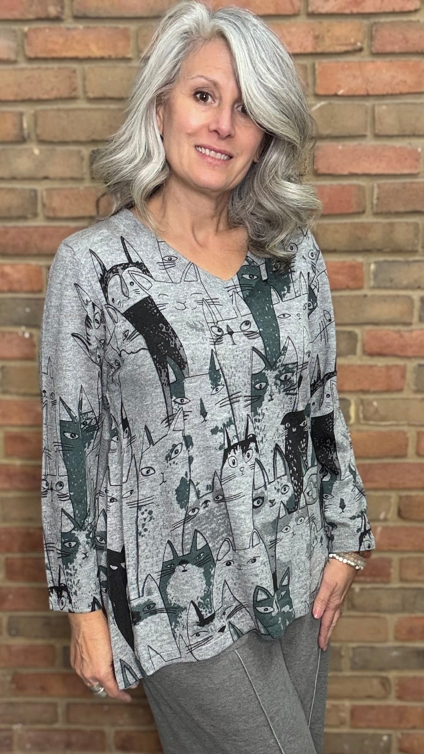 Woman wearing a patterned top and gray pants against a brick wall.