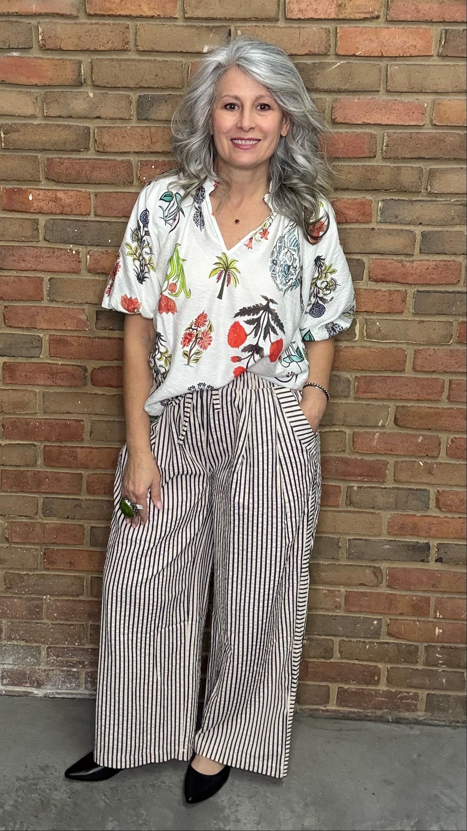 Woman in a floral blouse and Latte Wide Leg Pull On Pant standing against a brick wall.