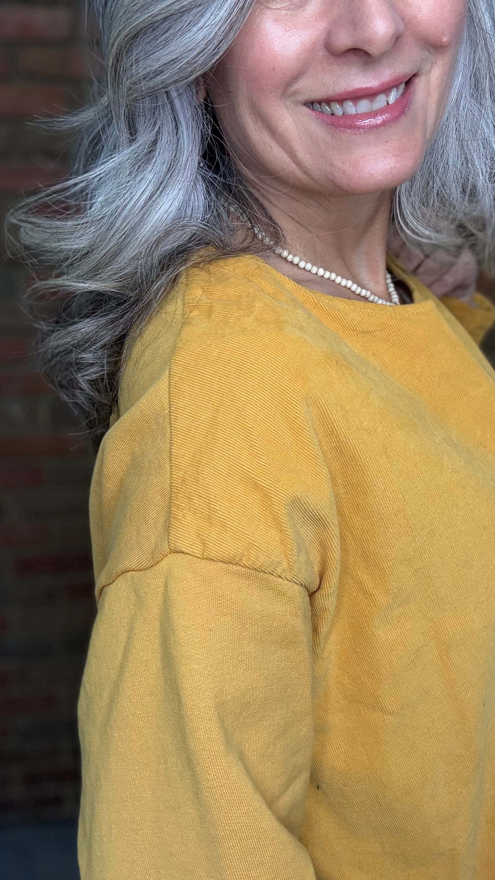 Woman wearing the Solid Mustard Puckered Pullover and the Upside Down Jeans standing in front of a brick wall.