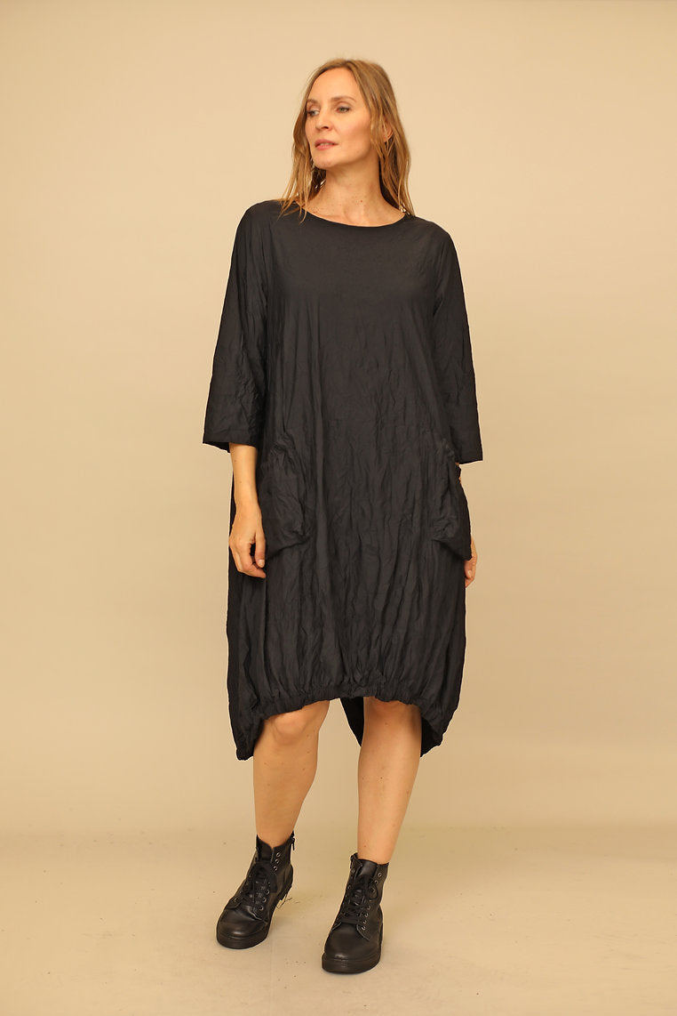 A model wearing a black ankle-length bubble dress with a round neckline, 3/4 sleeves, and front patch pockets, paired with black boots.