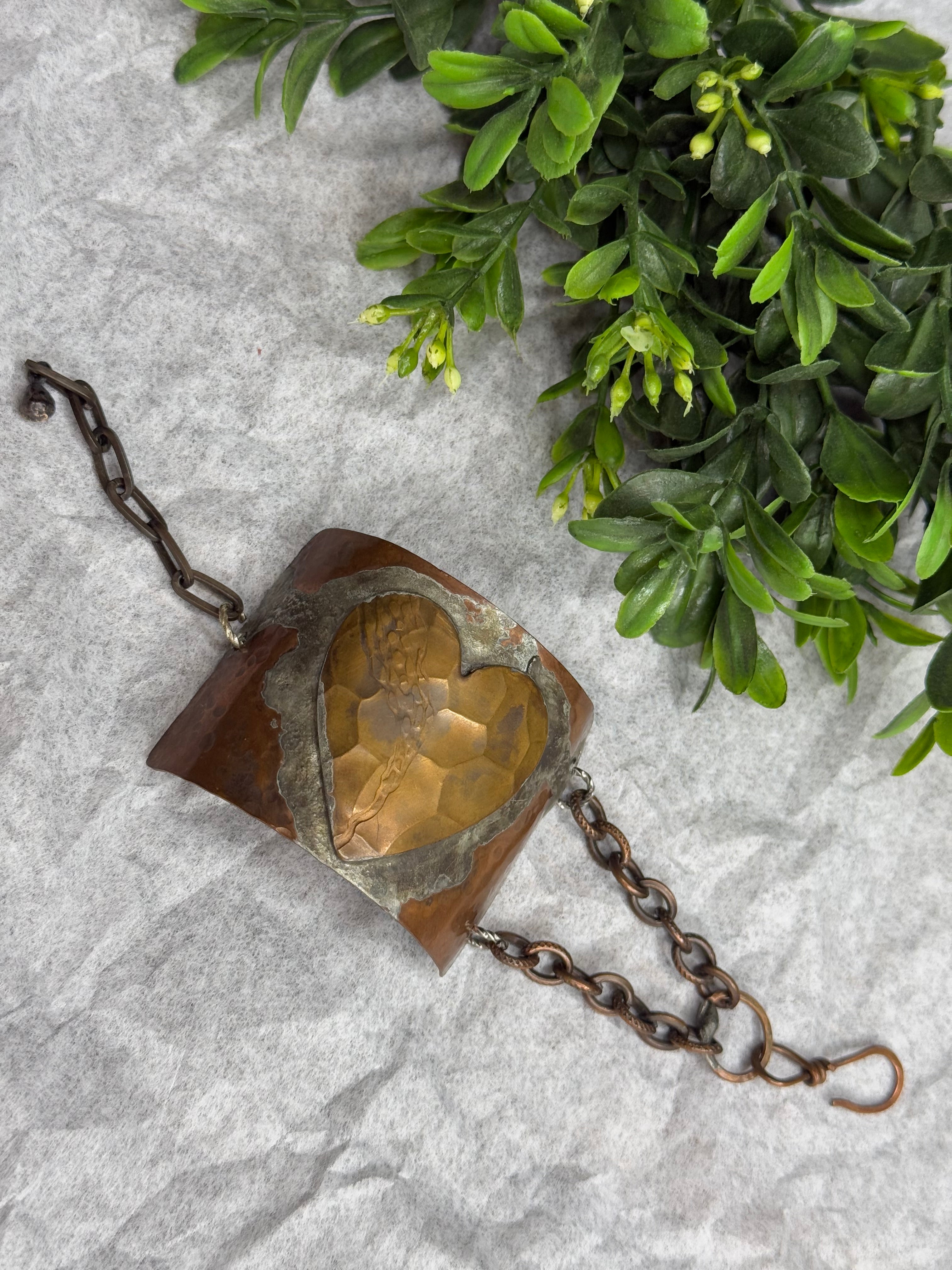 Bronze heart-shaped object with chain on a textured surface with greenery