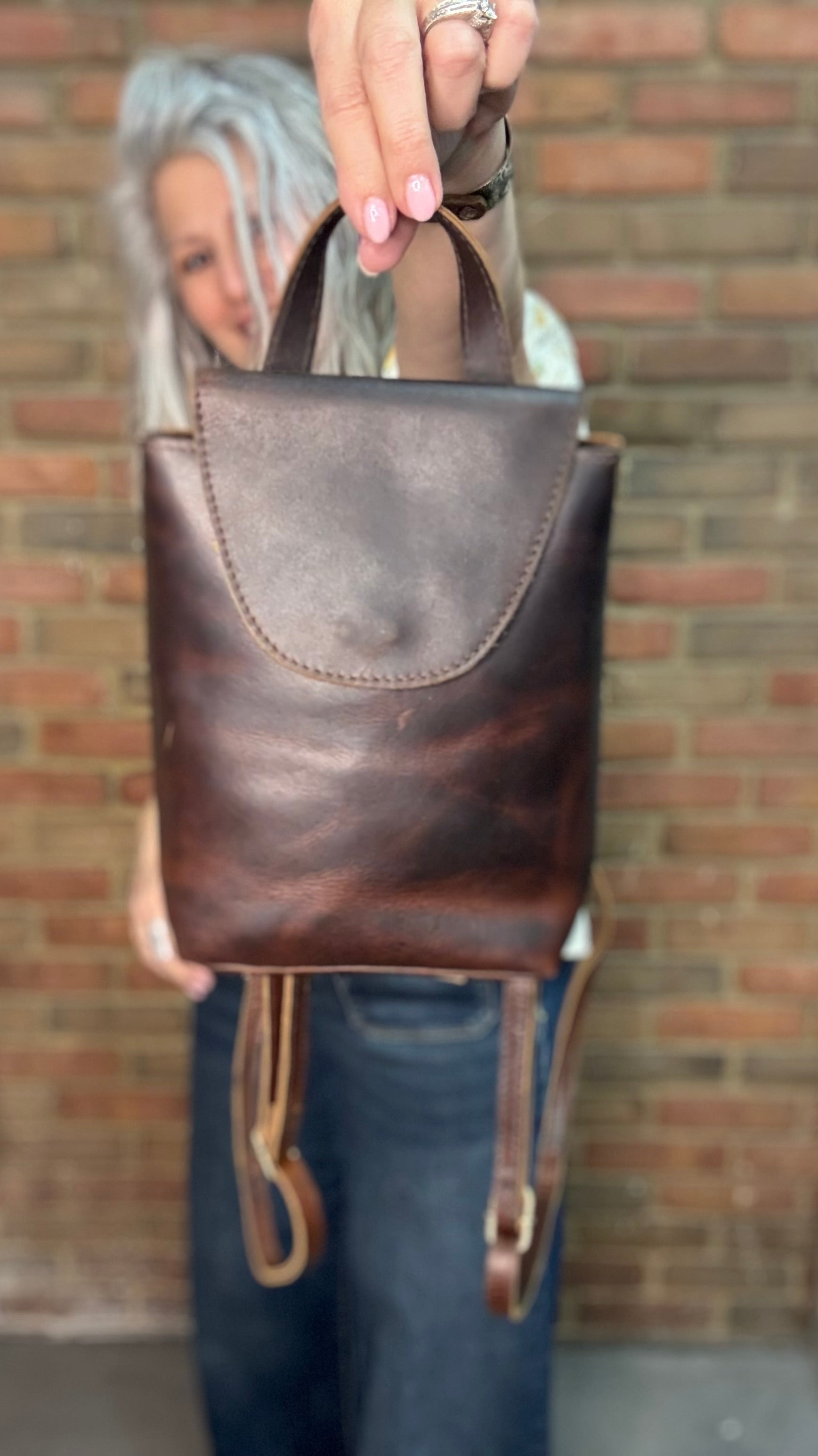Woman holding a Backpack style leather Handbag with a brick wall in background.