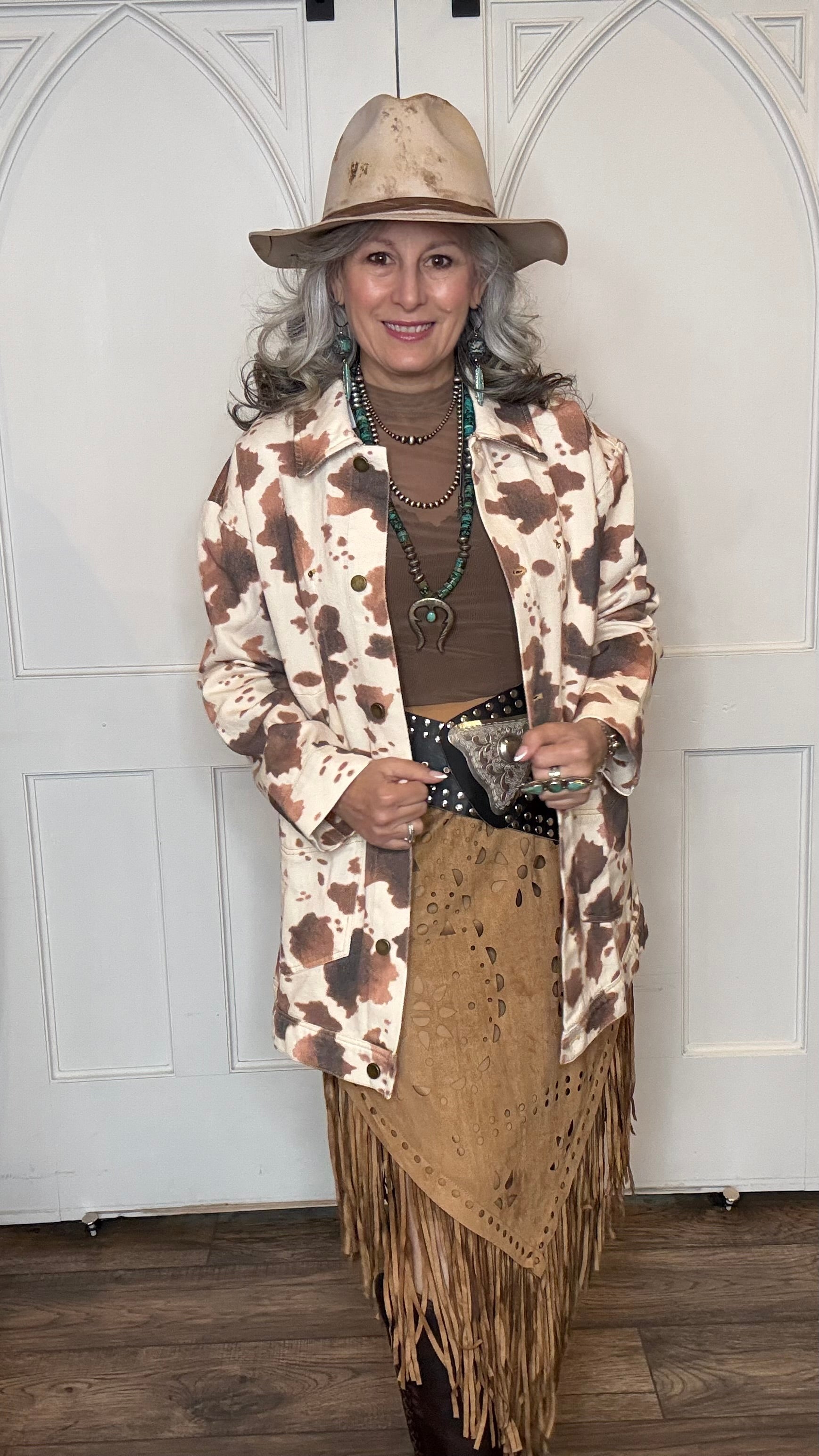 Woman wearing the Cow Print Denim Jacket, the Camel Faux Suede Fringe Skirt, topped the solid Mesh top in Brown and the Wide Triangle Buckle Belt. Shoes are the Lane Plain Jane Boots, and she is wearing the Pop&