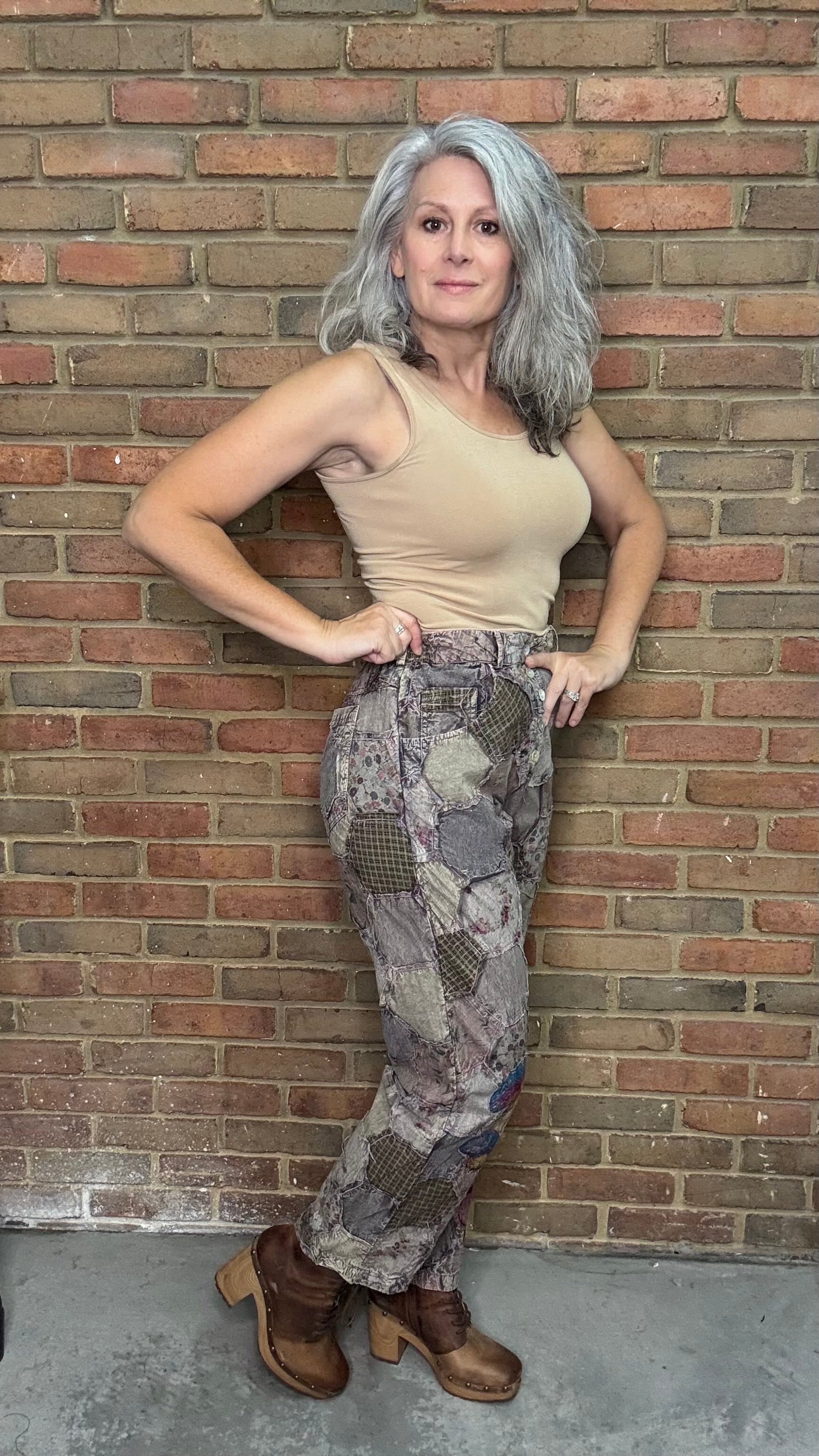 Woman posing against a brick wall wearing a beige top and patterned pants.
