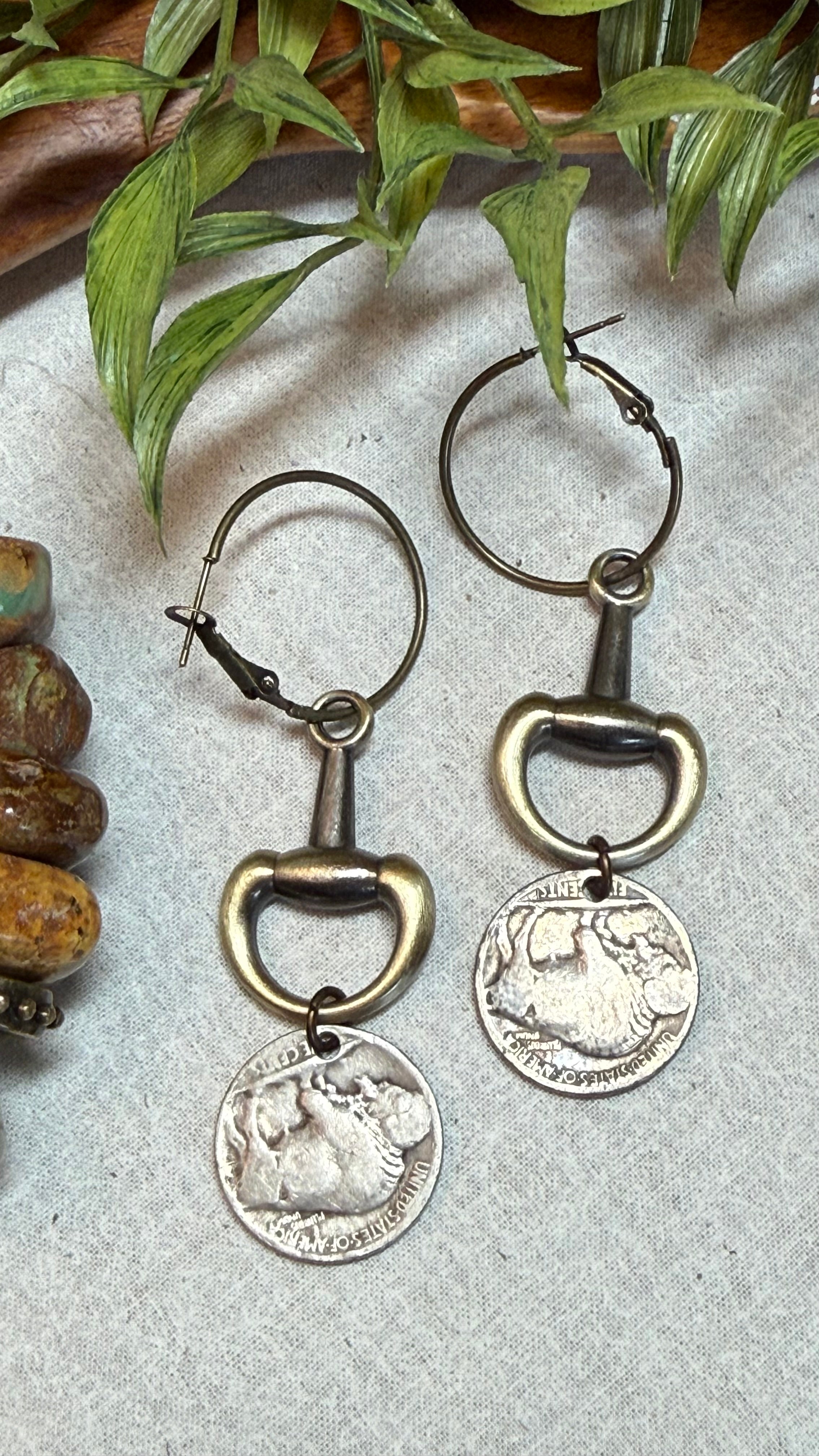 A pair of hoop earrings with antique bronze bottle opener charms and dangling silver coin pendants, displayed on a textured light gray surface with green leaves and brown stones as props.