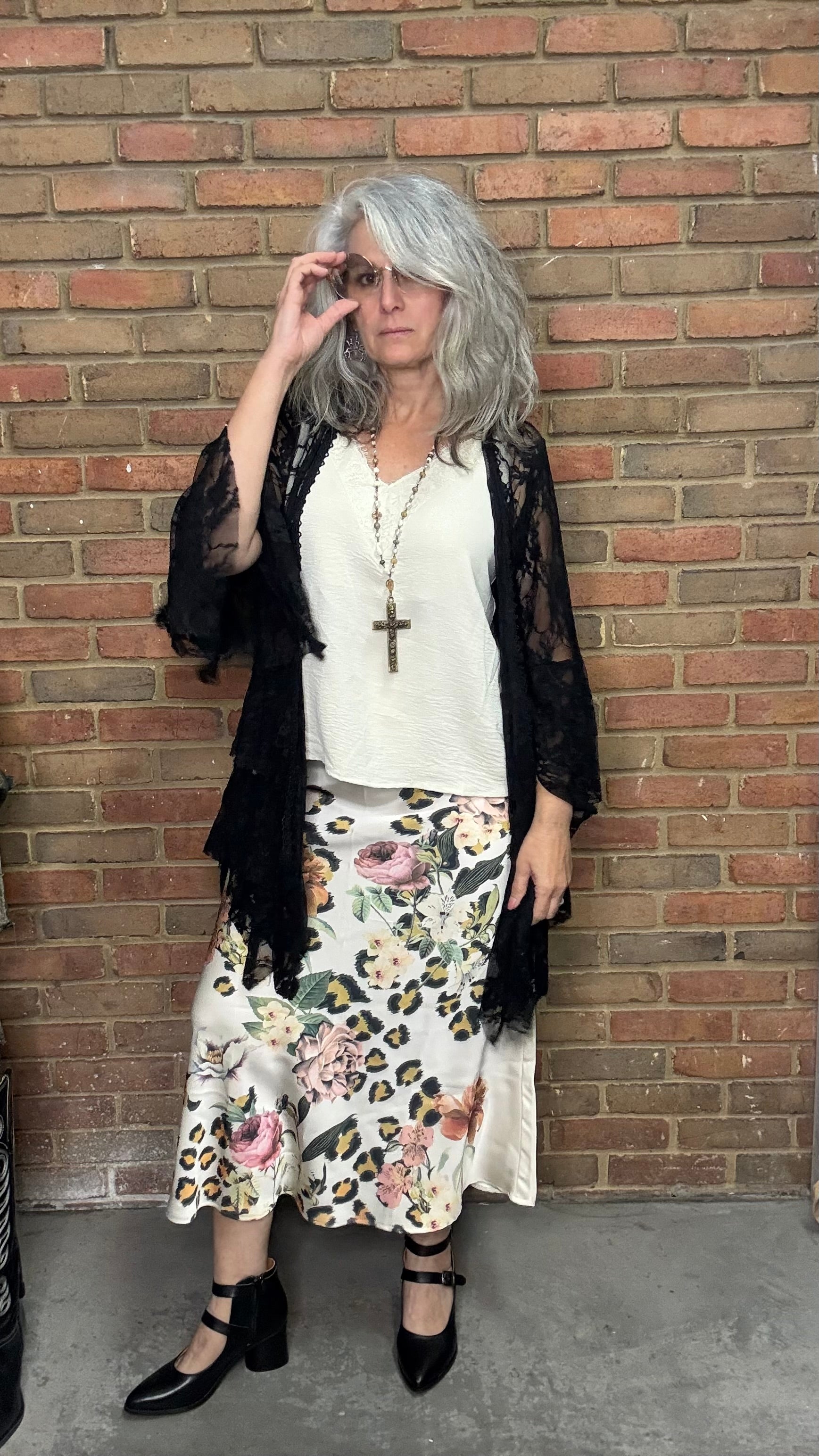 Woman in a floral skirt and black top standing against a brick wall.