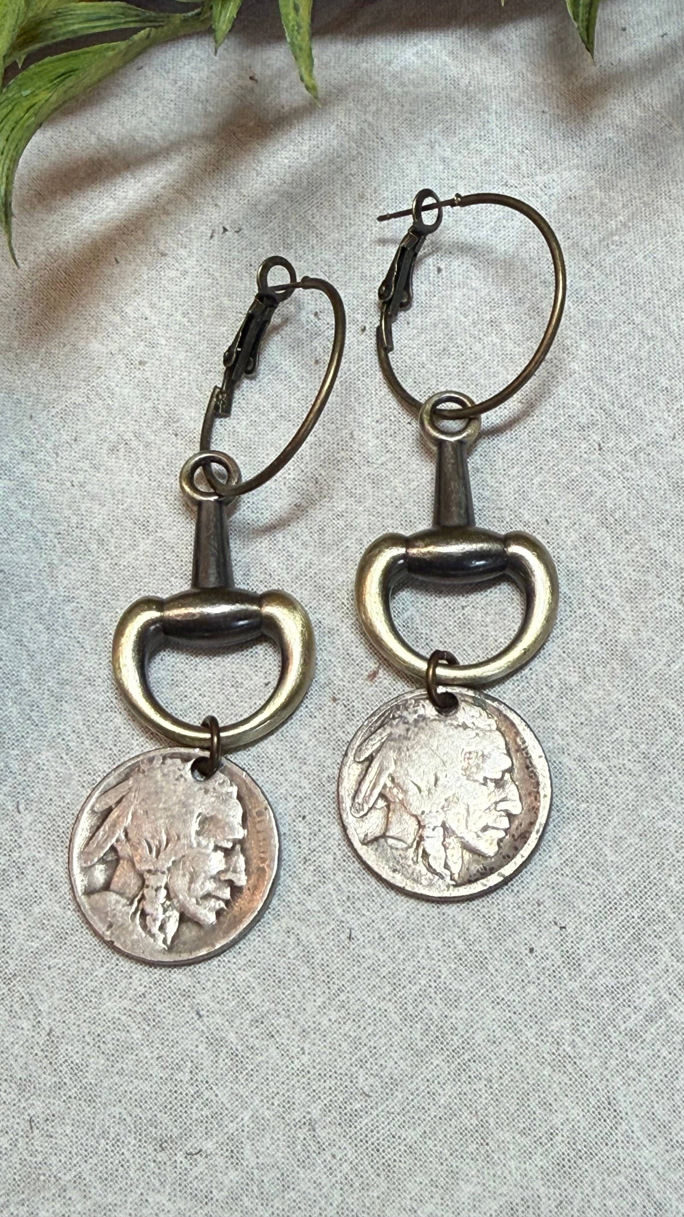 A pair of drop earrings made from antique bronze bottle opener rings and silver coin pendants, displayed on a textured light gray fabric background with green leaves in the corner.