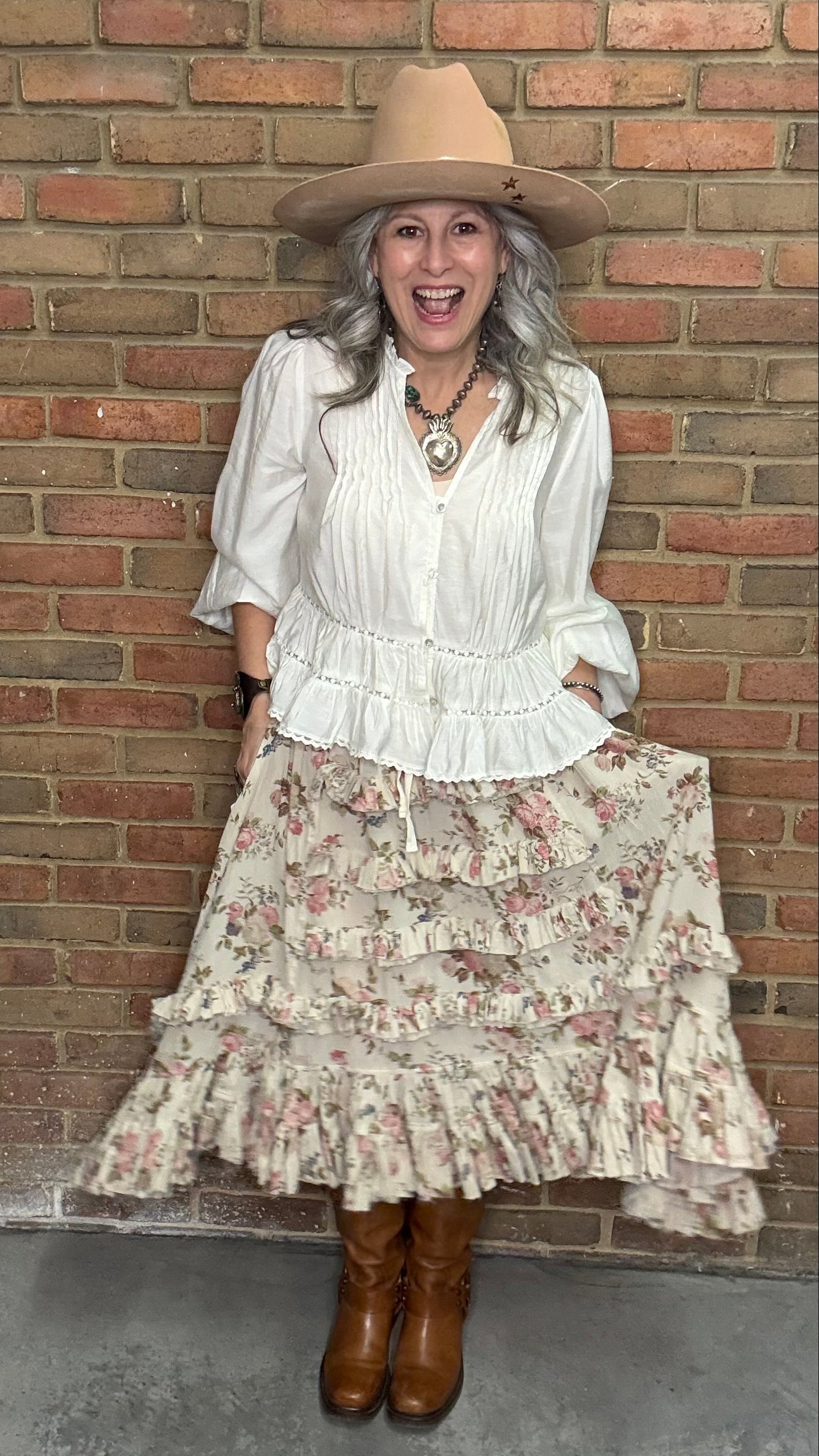 Woman in a White Pin-Tuck Lace Embellished shirt, blooming romance skirt, and Kate Harness Frye boots standing against a brick wall.