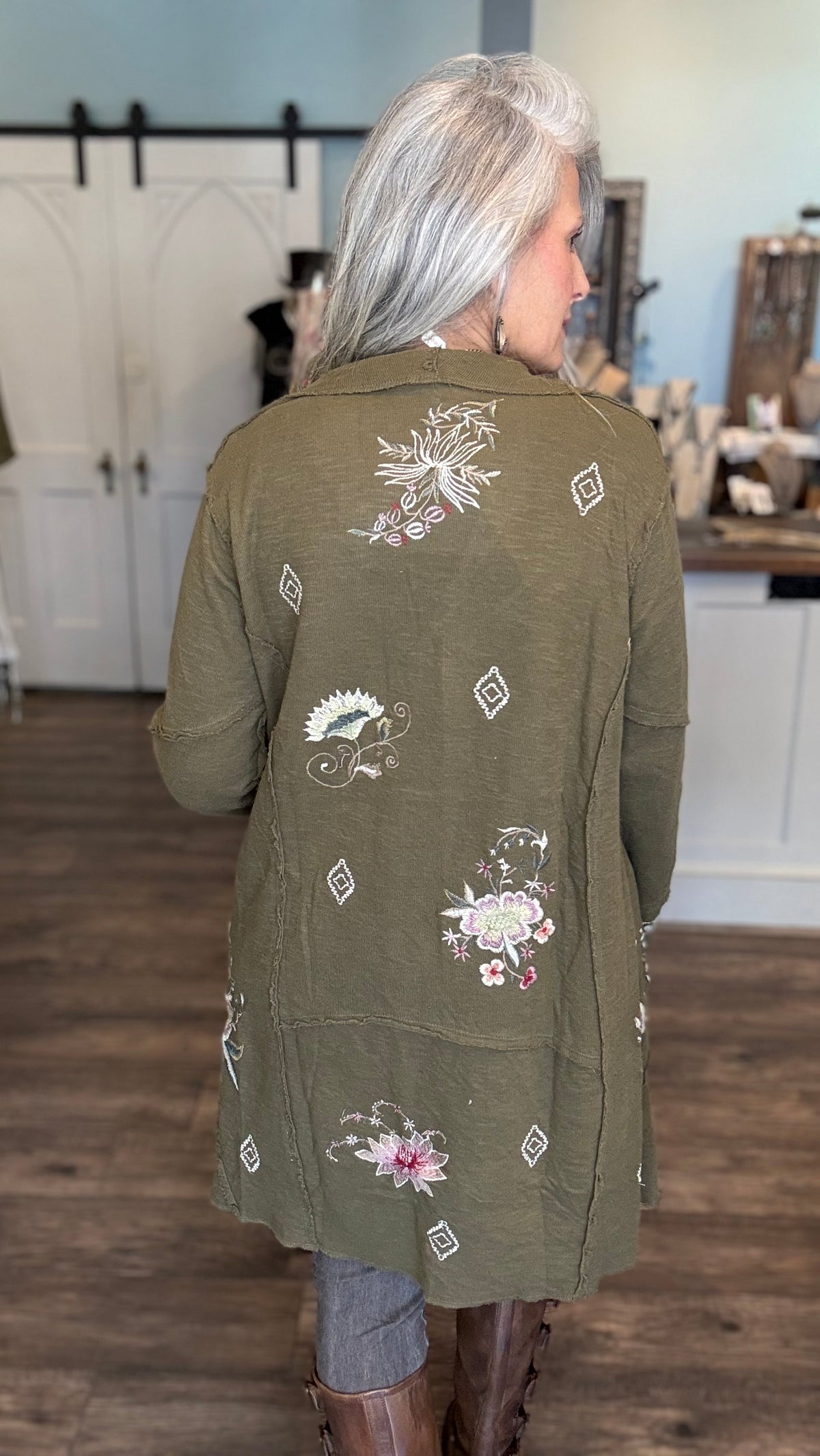 A woman viewed from behind wearing an olive green embroidered cardigan with floral and geometric patterns, standing in a rustic indoor shop setting with wood floors and white barn-style doors in the background.