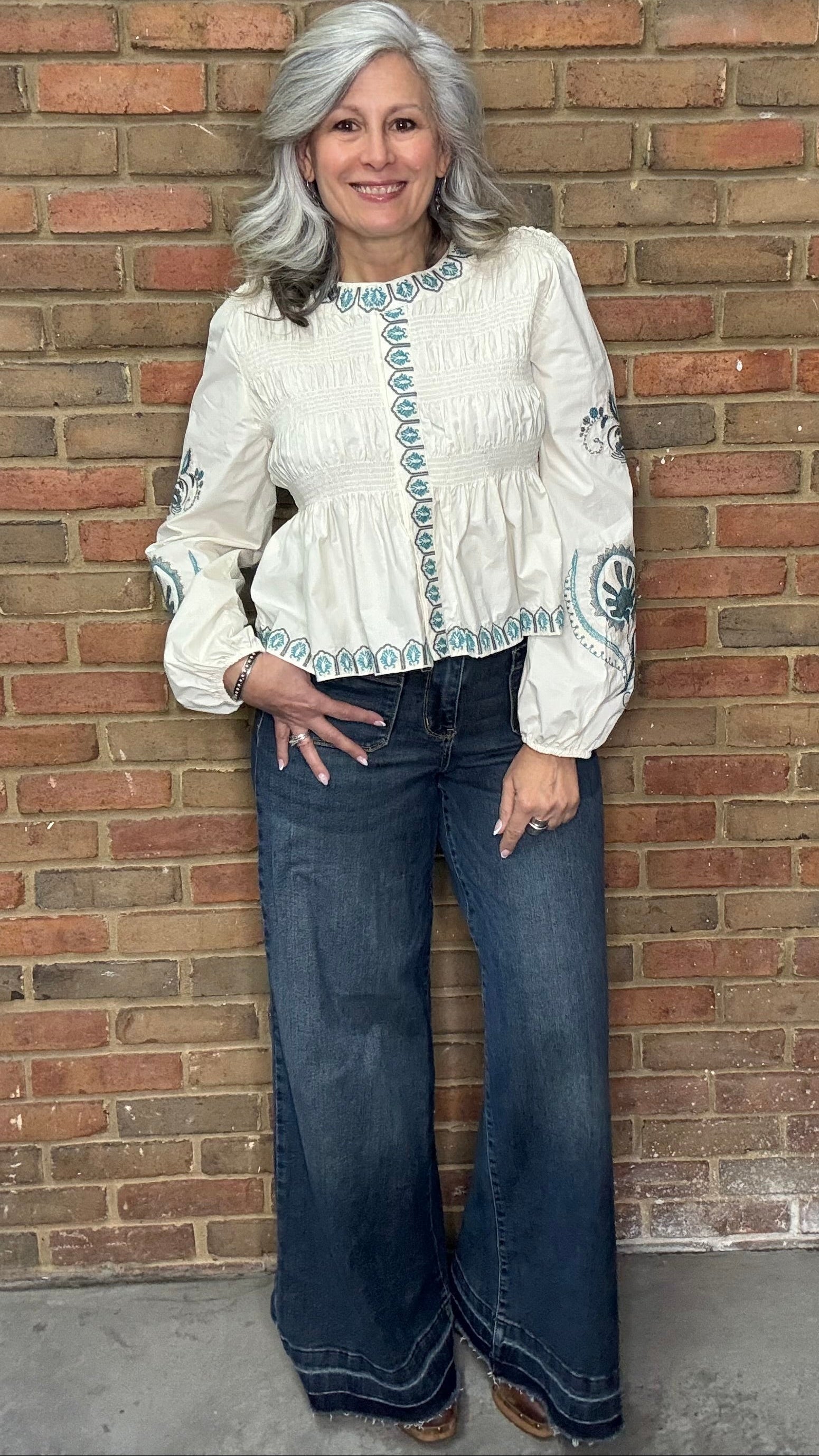 Woman wearing the Embroideried Smocked Peasant Top and Judy Blue Palazzo jeans against a brick wall.