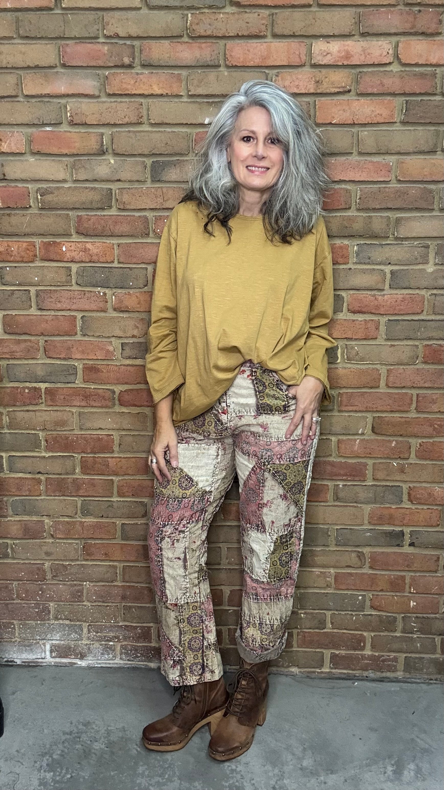 Woman standing against a brick wall wearing a mustard yellow top and patterned pants.