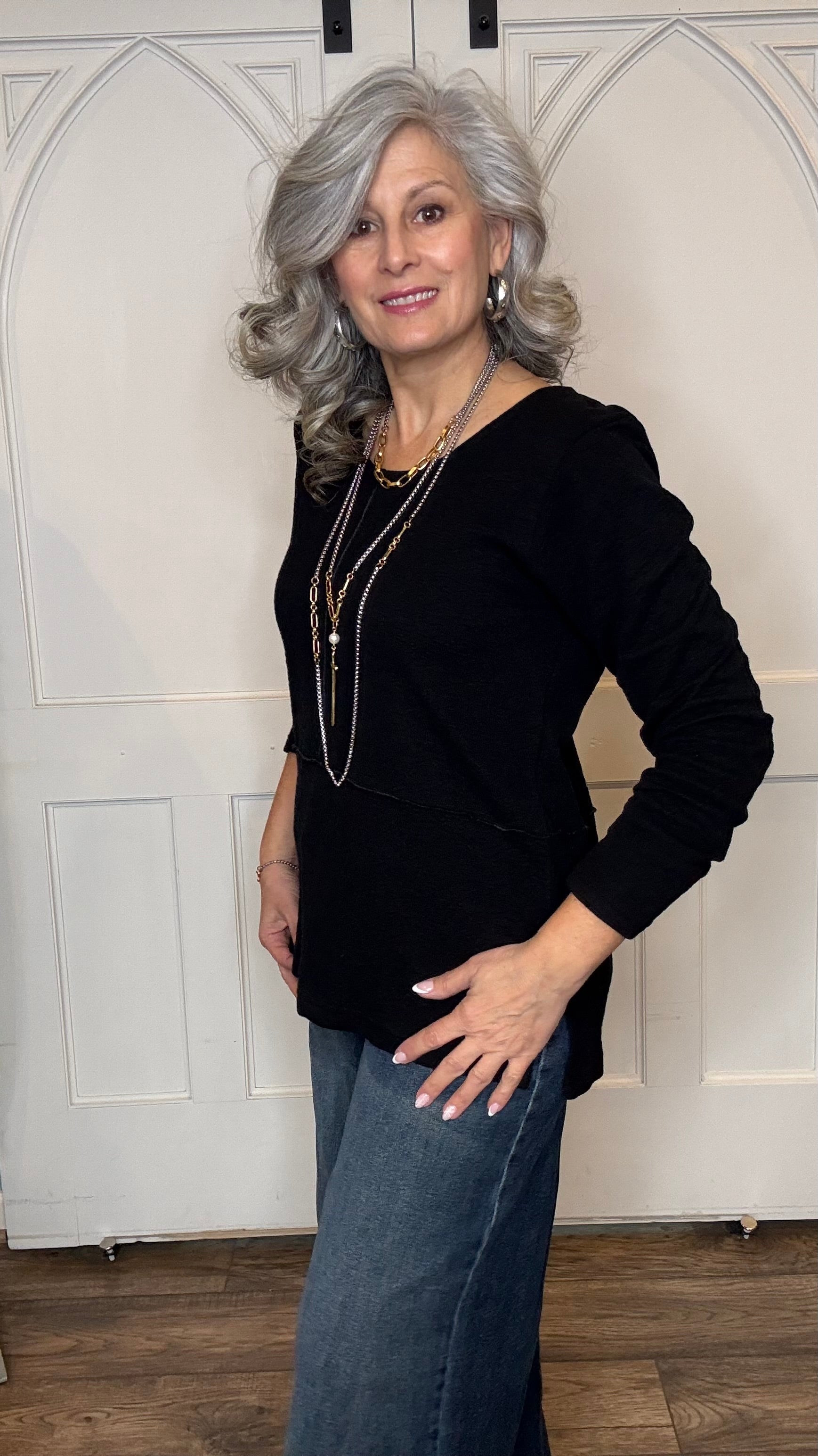 Woman wearing The Black Wanda Round Neck Top layered with the Silver Snake and Gold Paperclip Chain Necklace, the Silver Snake and Gold Paperclip Chain Choker Necklace and the Gold Cross w/Pearl Necklace paired with Judy Blue Palazzo jeans indoors against a white paneled wall.
