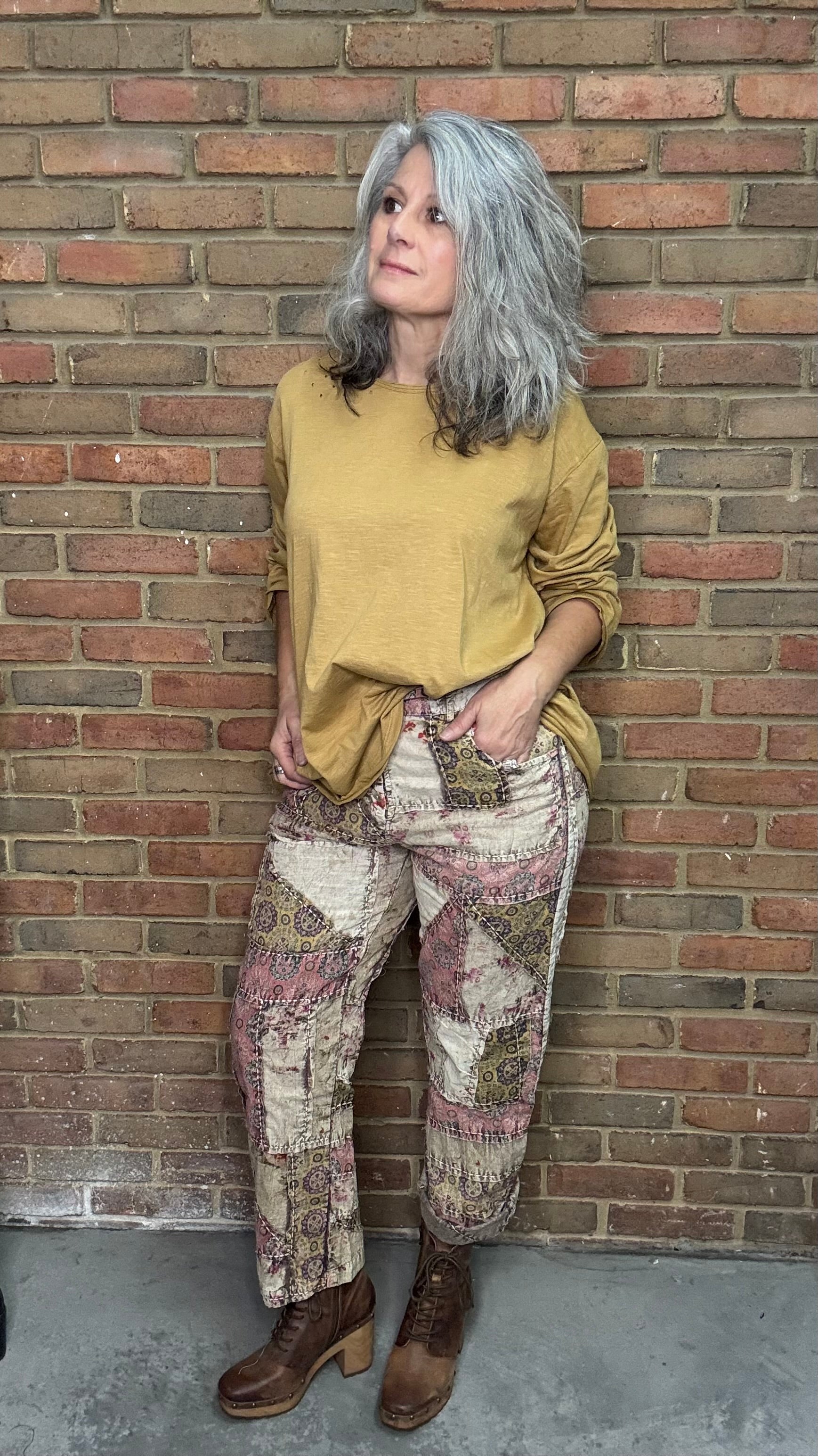 Person wearing a mustard yellow sweater and patterned pants leaning against a brick wall.