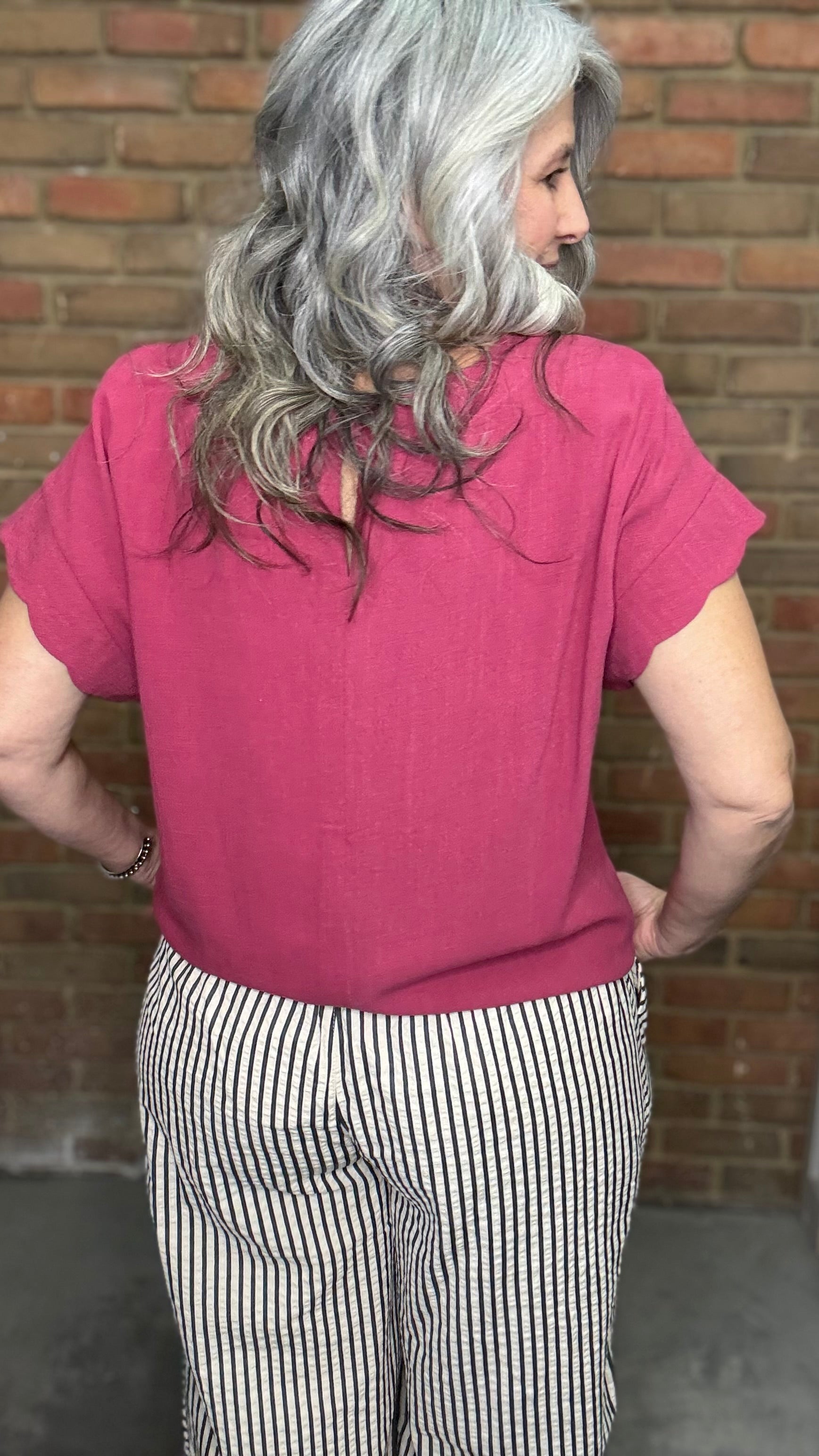 Woman wearing Scalloped Sleeve Tie Hem Top - Berry and striped pants standing against a brick wall.