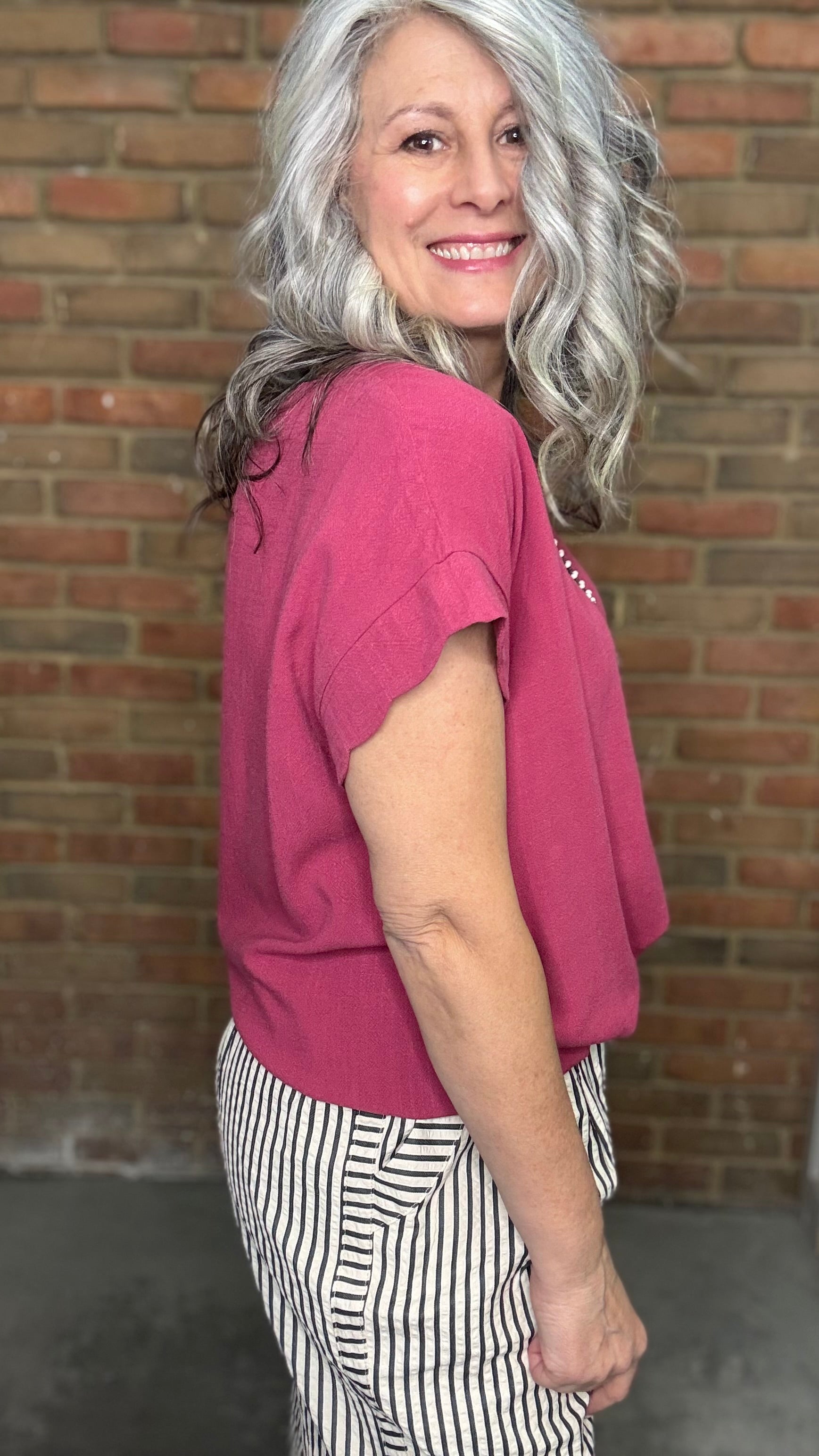 Woman wearing Scalloped Sleeve Tie Hem Top - Berry and striped pants standing against a brick wall.