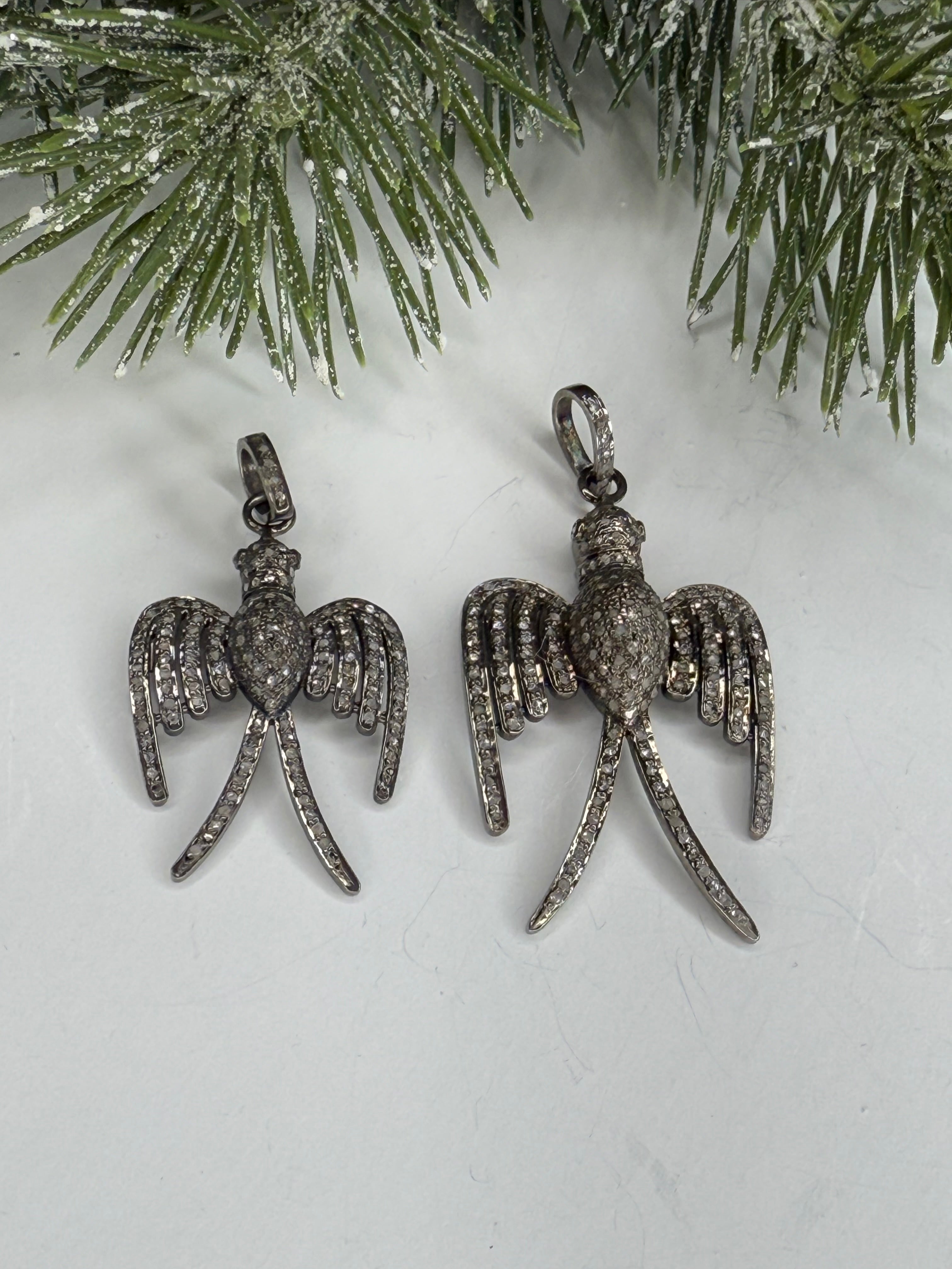 Bird shaped pendants with pave diamond detailing on a white surface with a green pine branch.