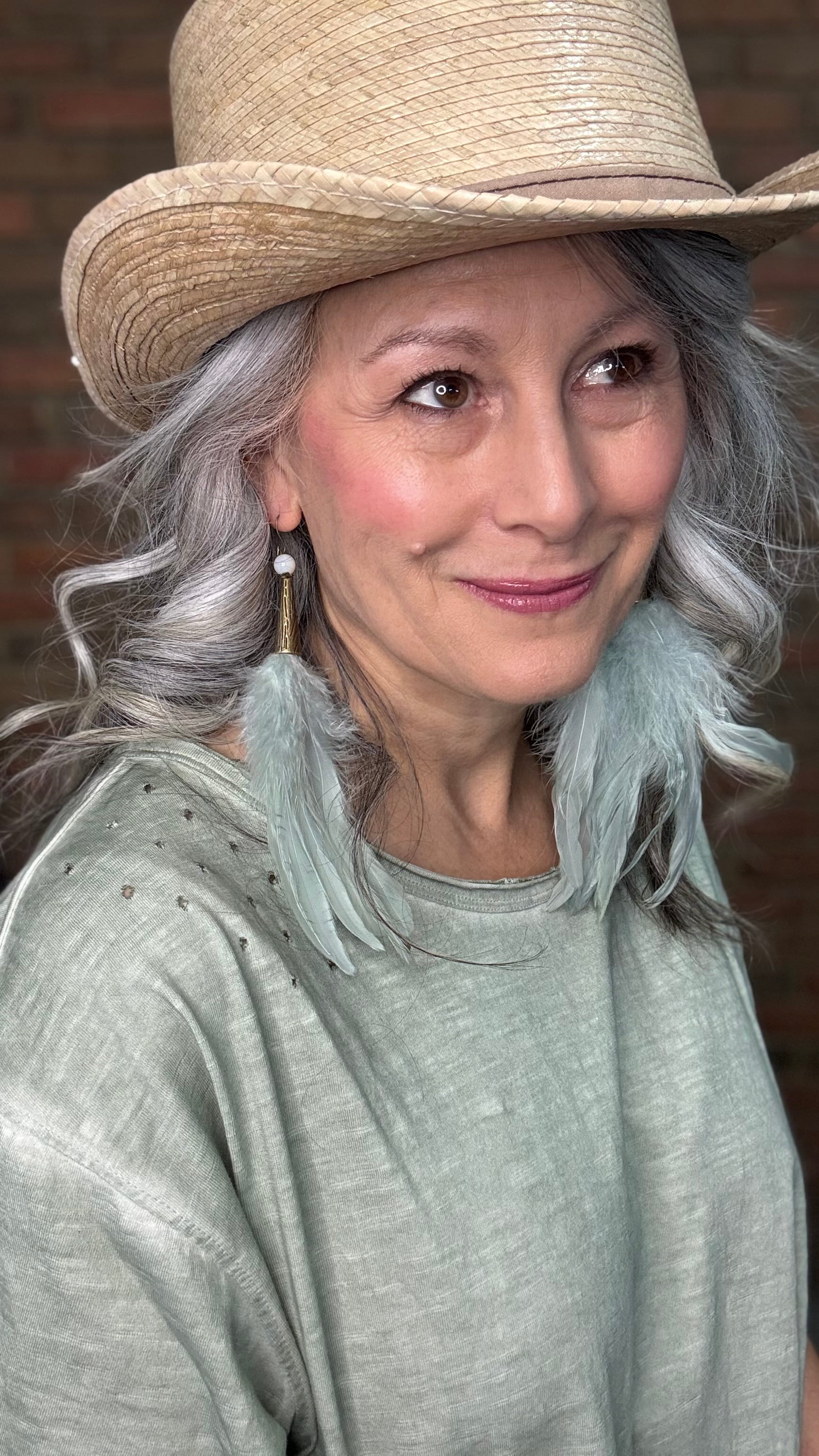 Woman wearing a straw hat with Sage Feather Earrings and a gray top against a brick wall.