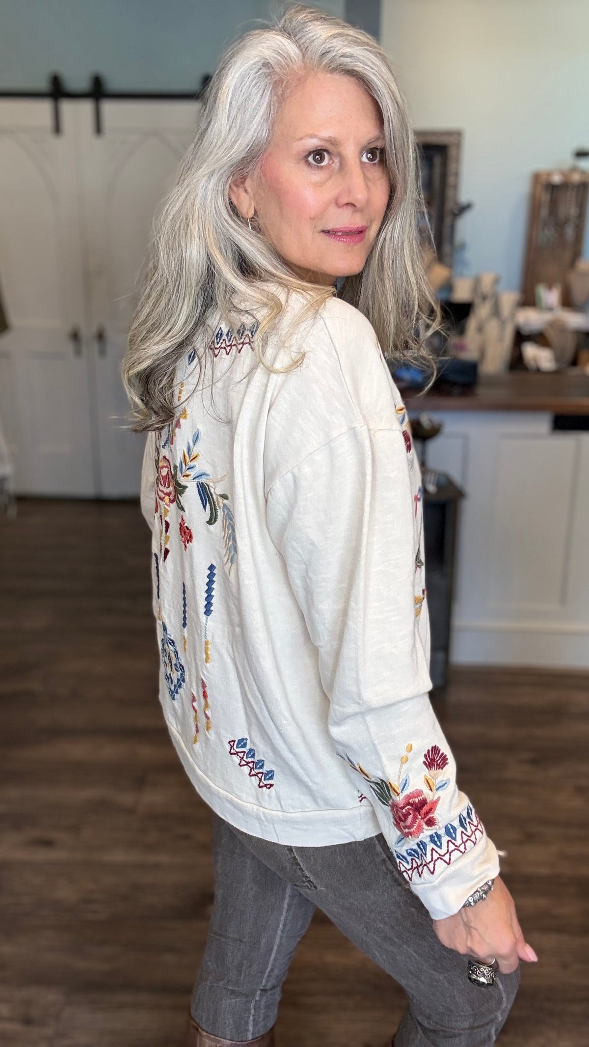 A woman with long gray hair is shown from the back wearing a cream-colored embroidered jacket with colorful floral and geometric patterns, paired with gray pants in an indoor home setting.