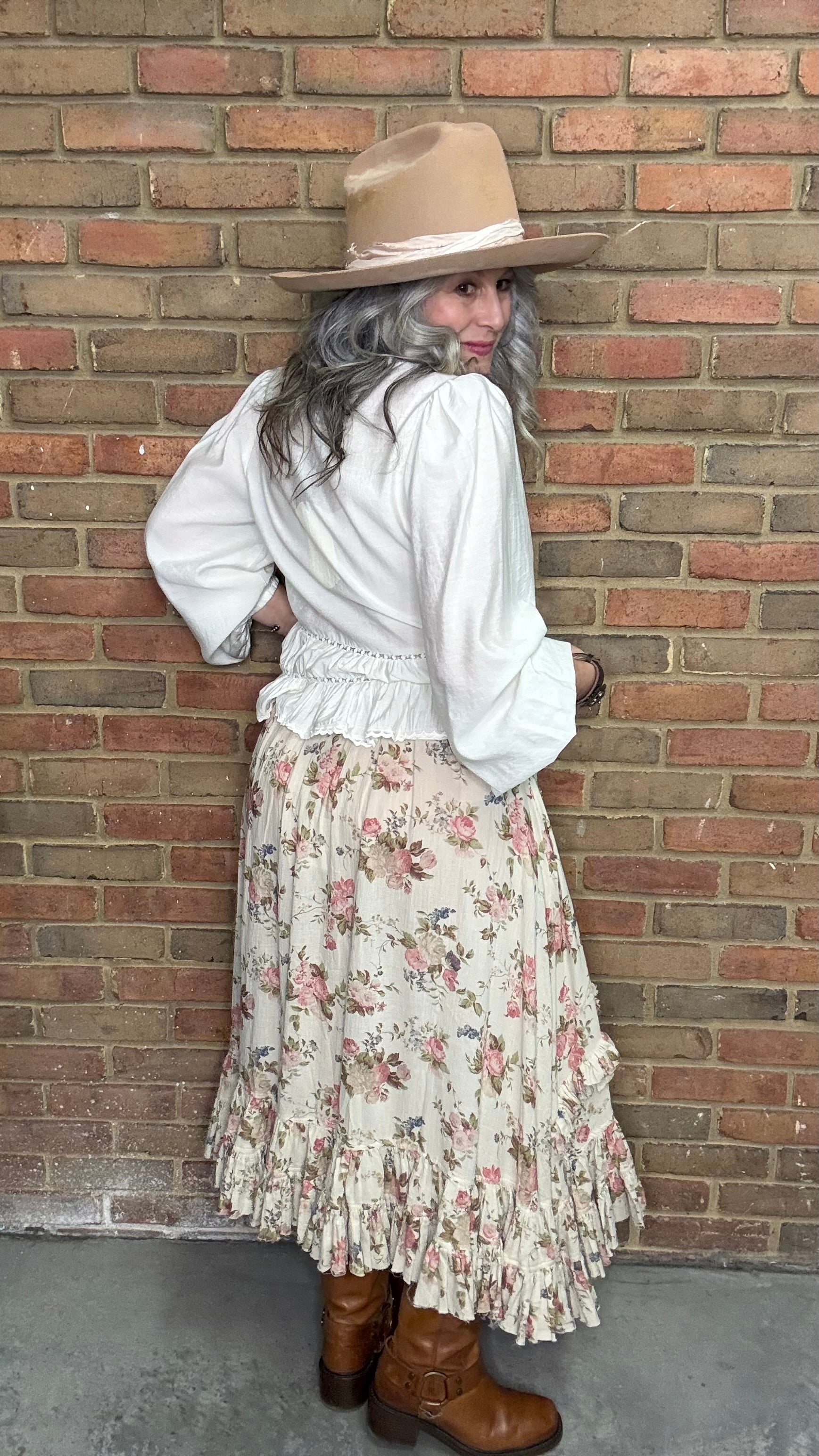 Woman in a White Pin-Tuck Lace Embellished shirt, blooming romance skirt, and Kate Harness Frye boots standing against a brick wall.
