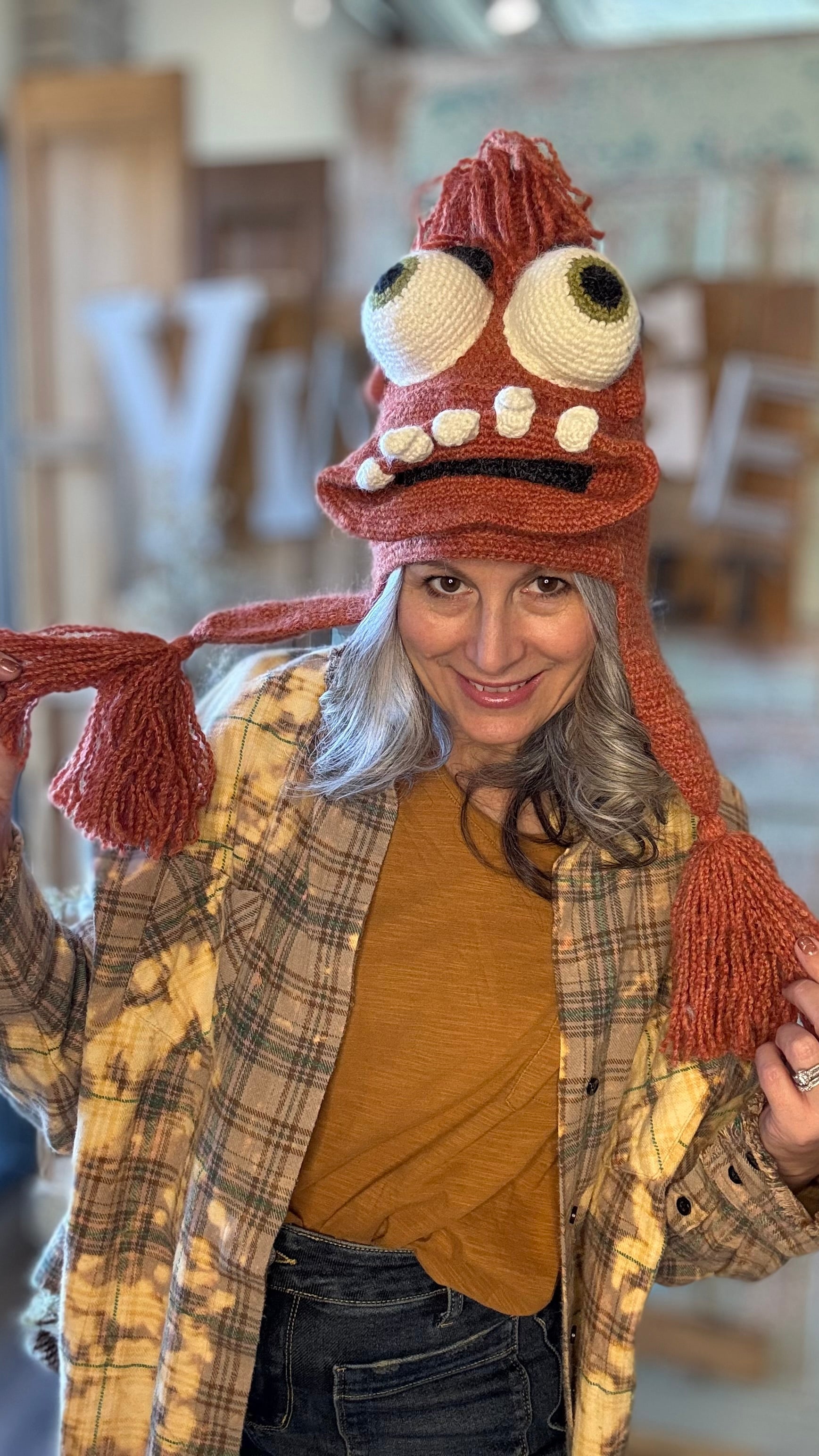 Person wearing a unique, eye-shaped hat with a plaid jacket and mustard shirt indoors.