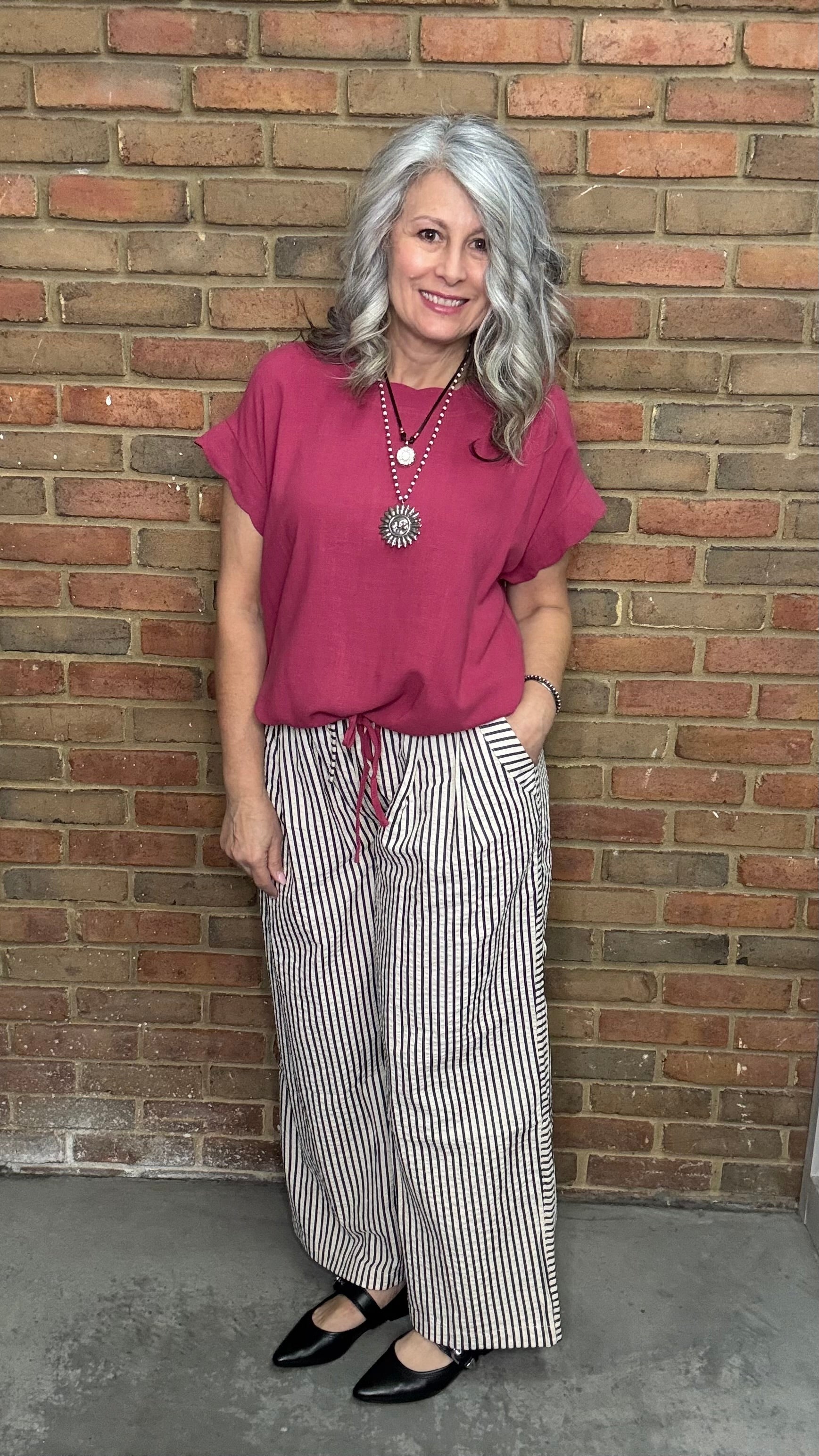 Woman wearing Scalloped Sleeve Tie Hem Top - Berry and striped pants standing against a brick wall.