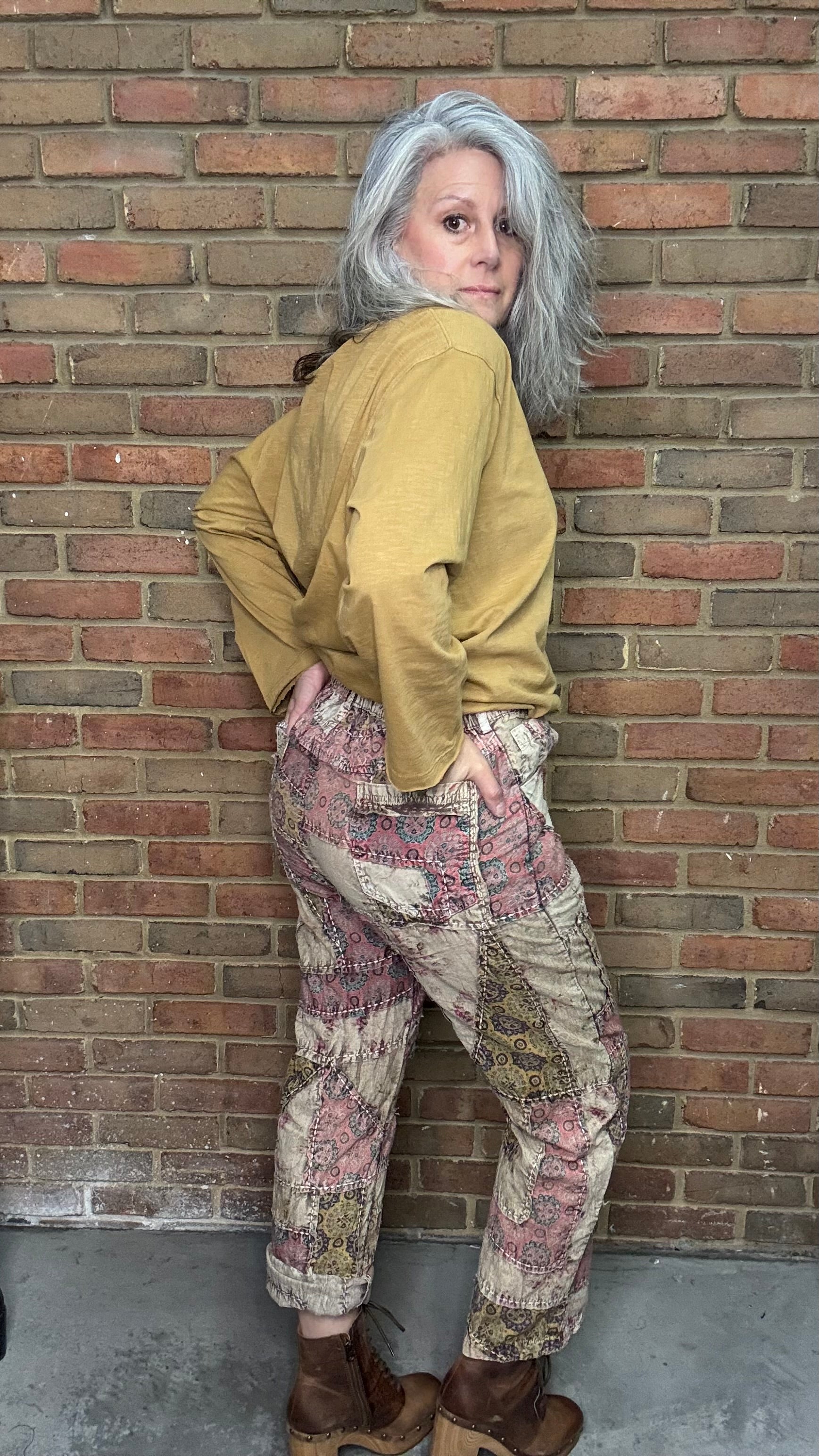 Person wearing a mustard jacket and patterned pants leaning against a brick wall.
