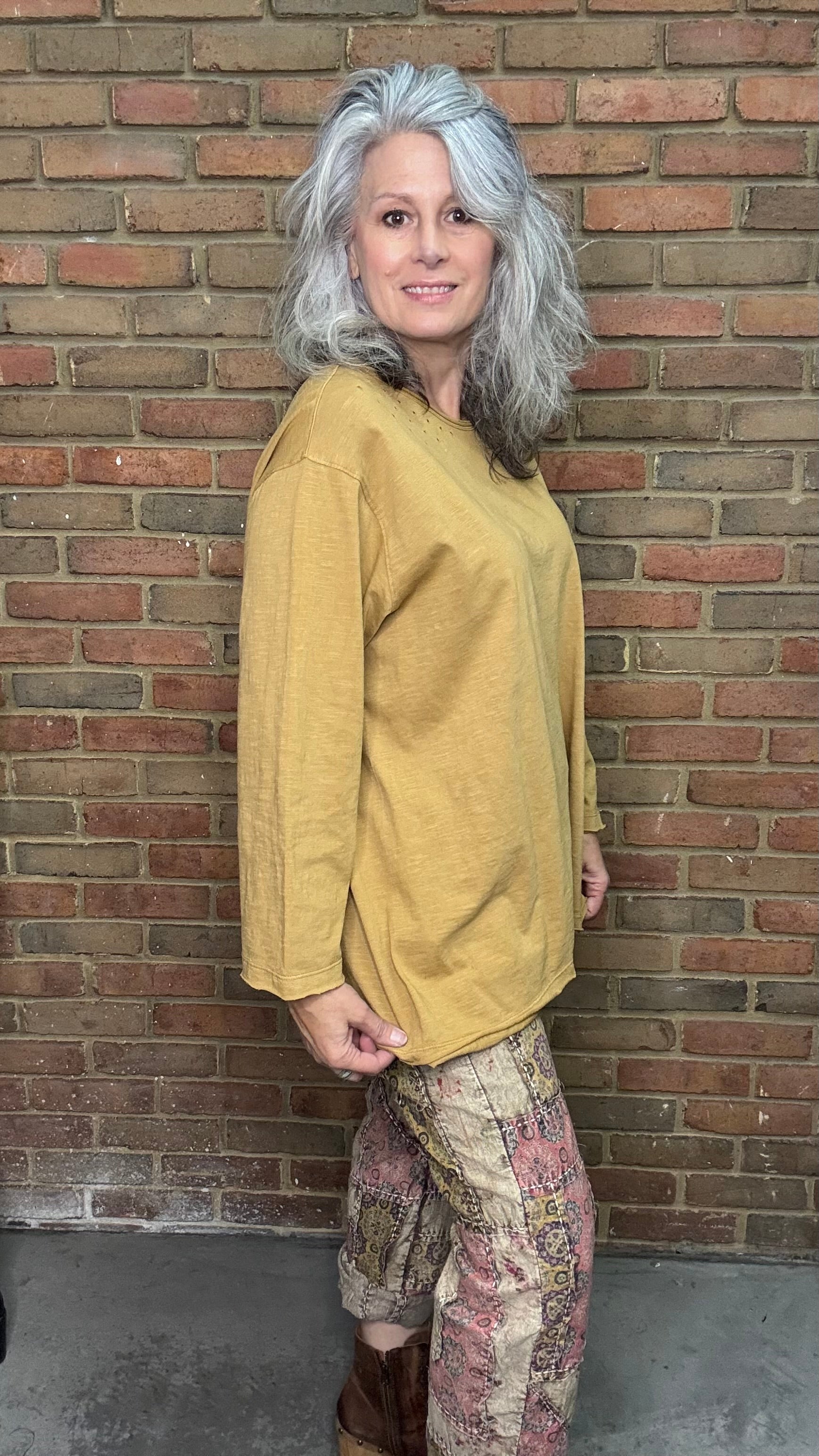 Person wearing a mustard yellow sweater and patterned pants standing against a brick wall.
