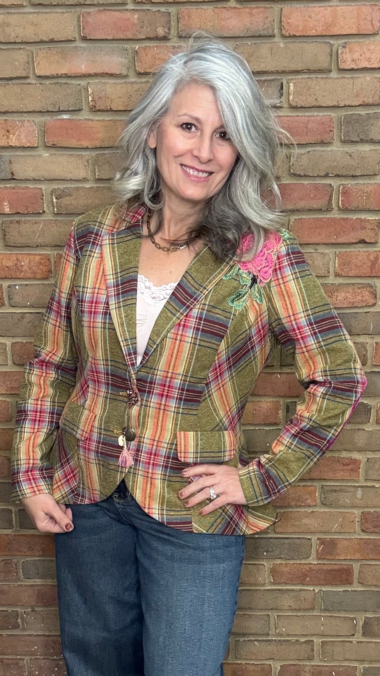 Woman wearing a plaid jacket and jeans standing against a brick wall.