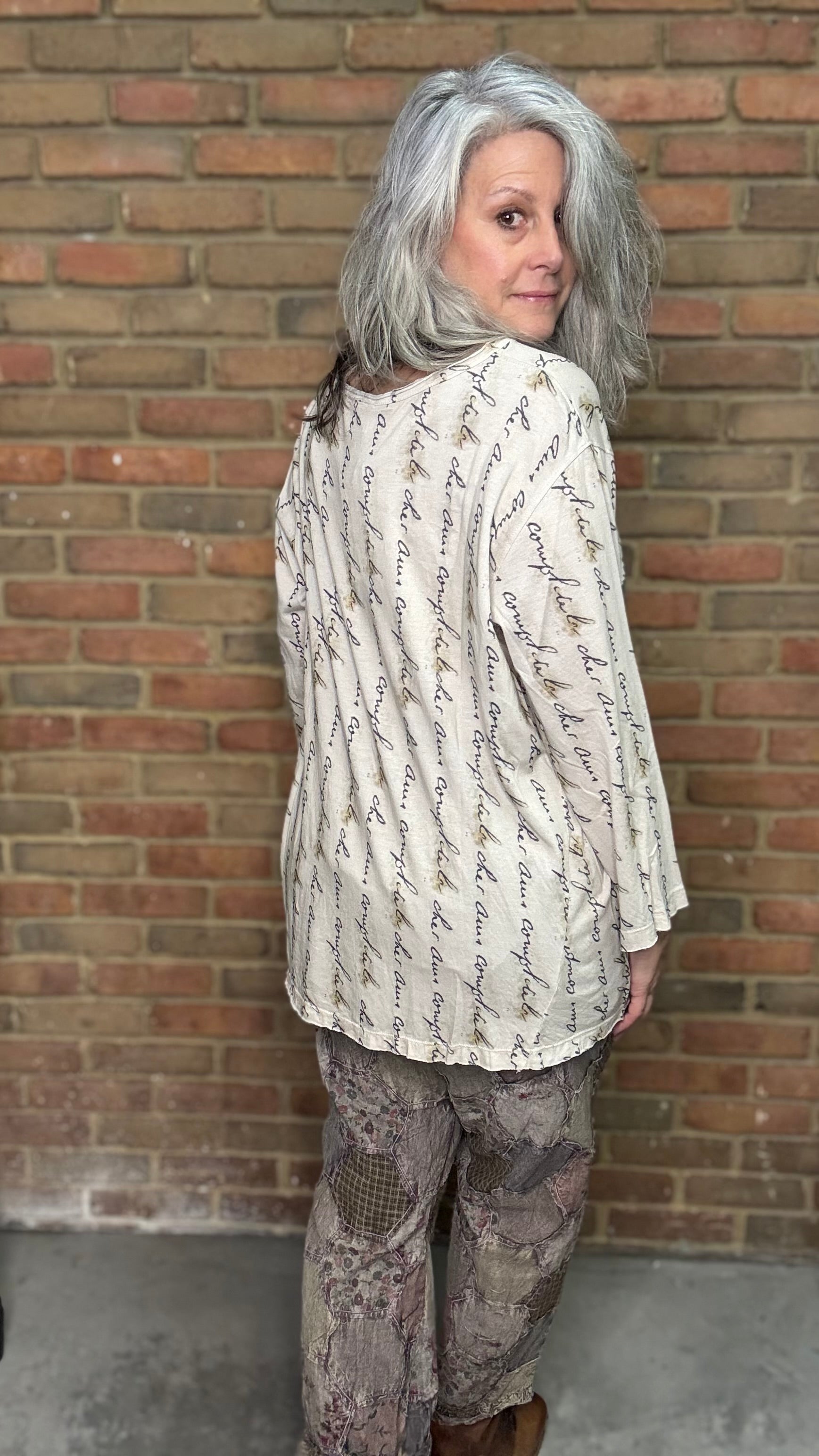 Person wearing a patterned shirt and pants standing against a brick wall.