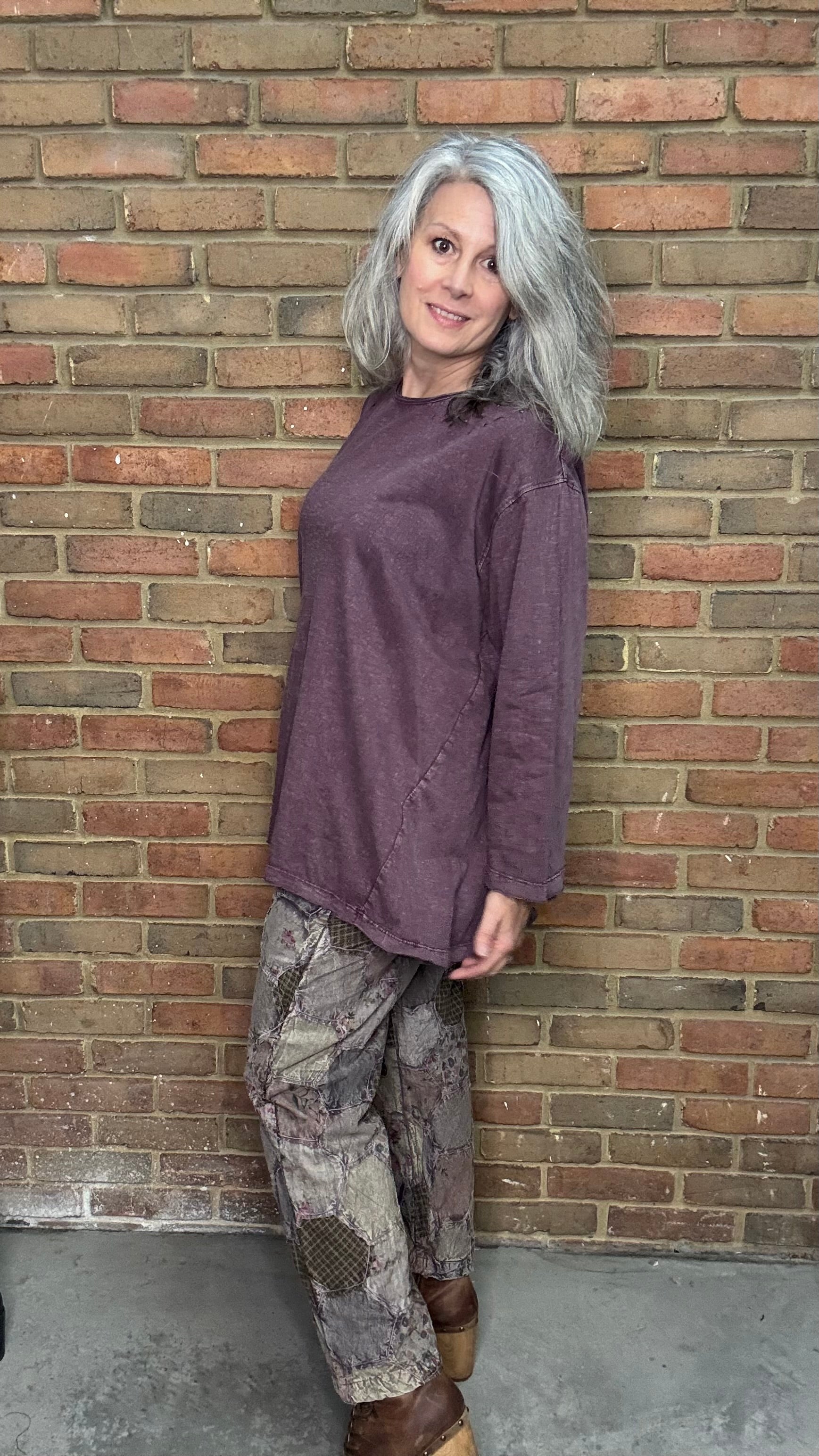 Woman wearing a purple shirt and patterned pants leaning against a brick wall.