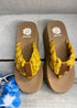 A pair of yellow platform sandals with braided straps and a square toe. The sandals are placed on a wooden surface with a signature yellow box logo visible.