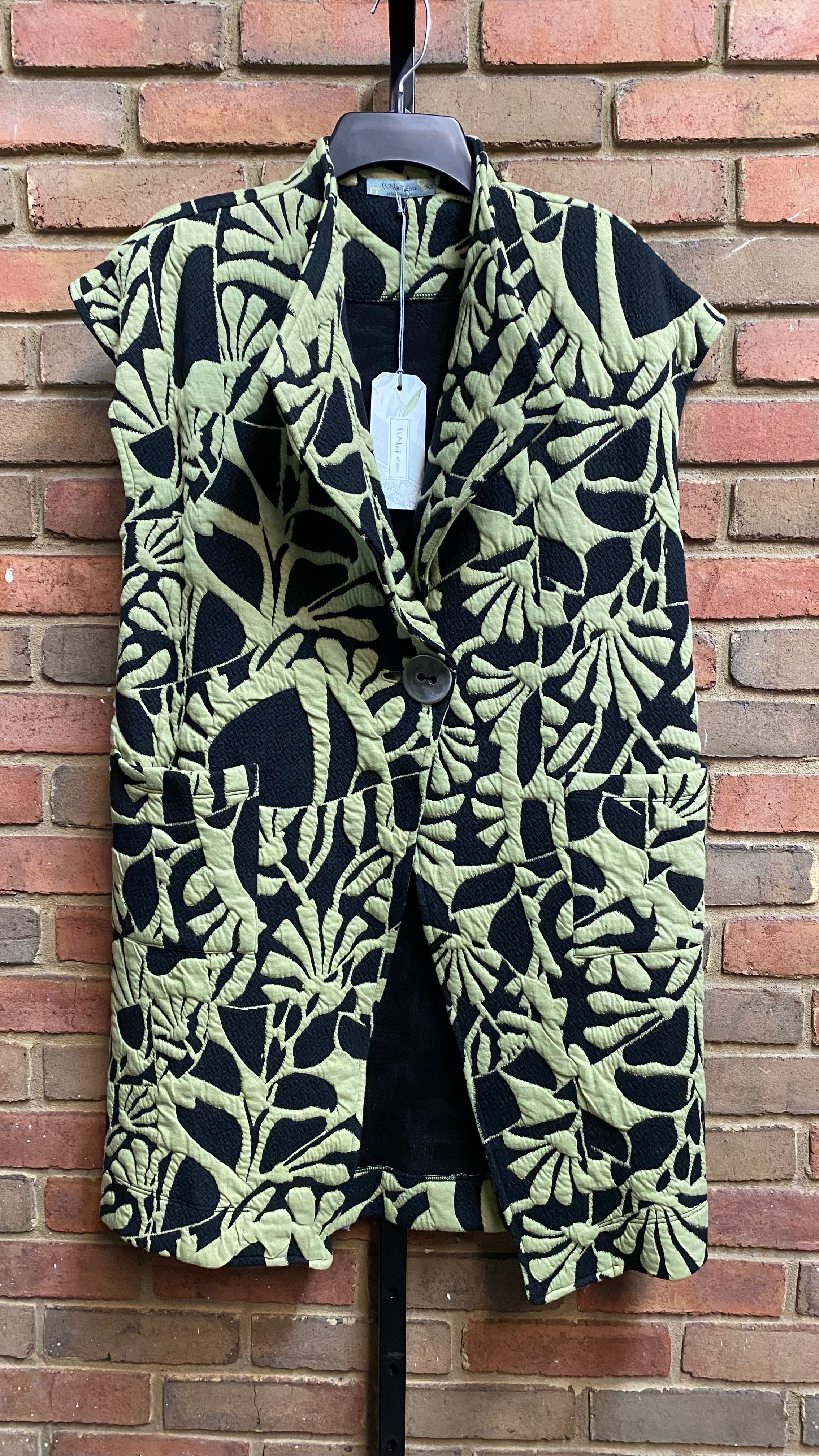 A black and green floral patterned vest with a wide lapel and a single large button, displayed on a hanger against a brick wall background.