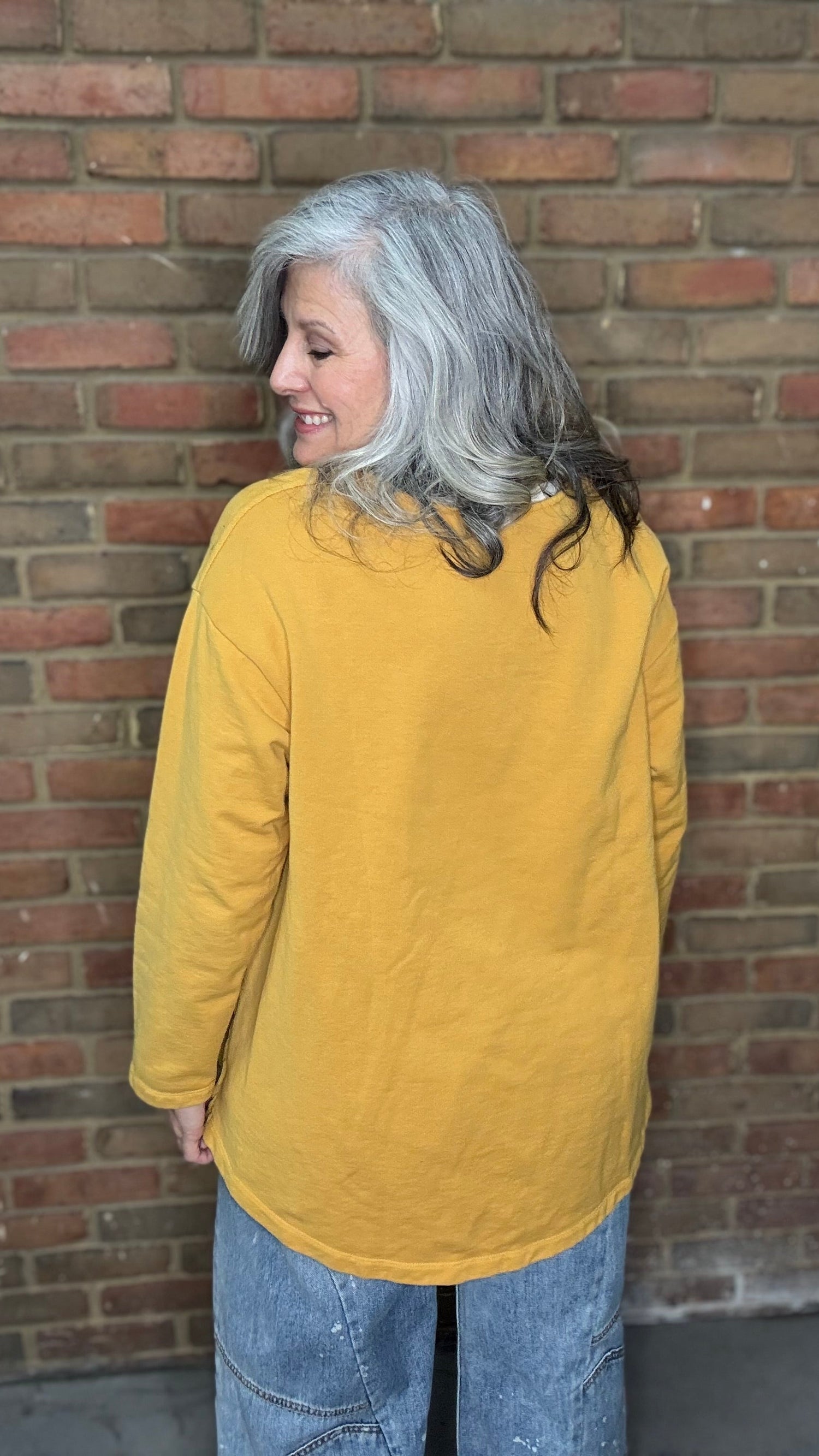 Woman wearing the Solid Mustard Puckered Pullover  and the Upside Down Jeans standing in front of a brick wall.
