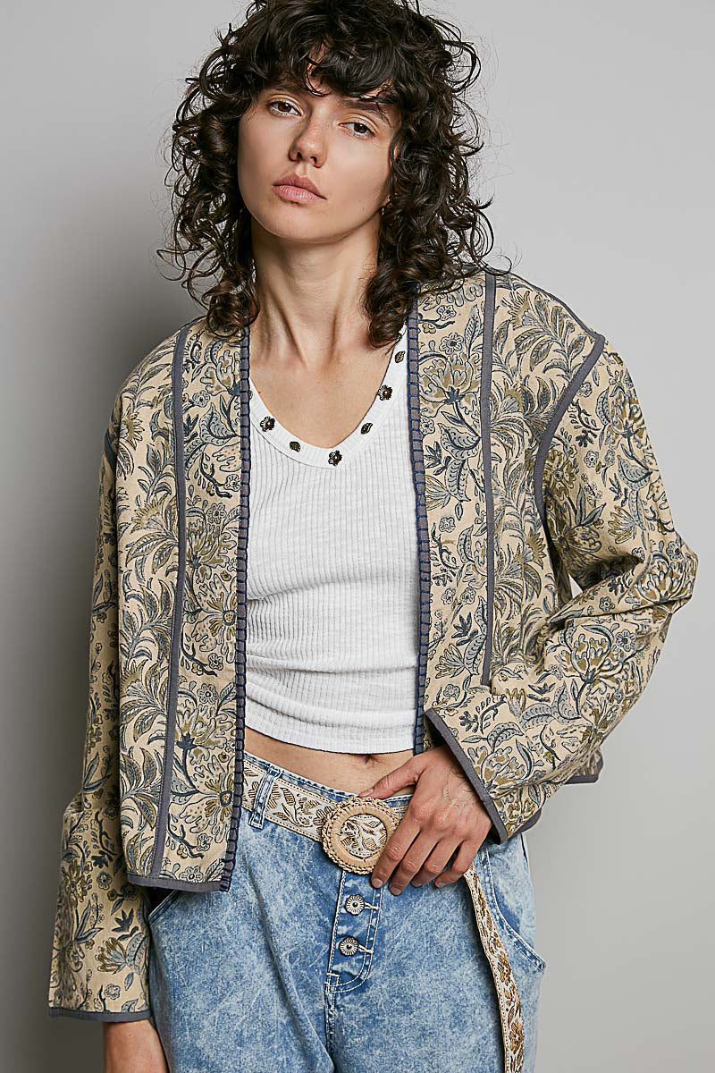 A model wearing a bohemian-style open-front jacket with a beige and brown paisley pattern, styled over a white ribbed crop top and blue jeans with a large decorative belt buckle.