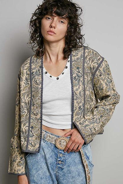 A model wearing a bohemian-style open-front jacket with a beige and brown paisley pattern, styled over a white ribbed crop top and blue jeans with a large decorative belt buckle.