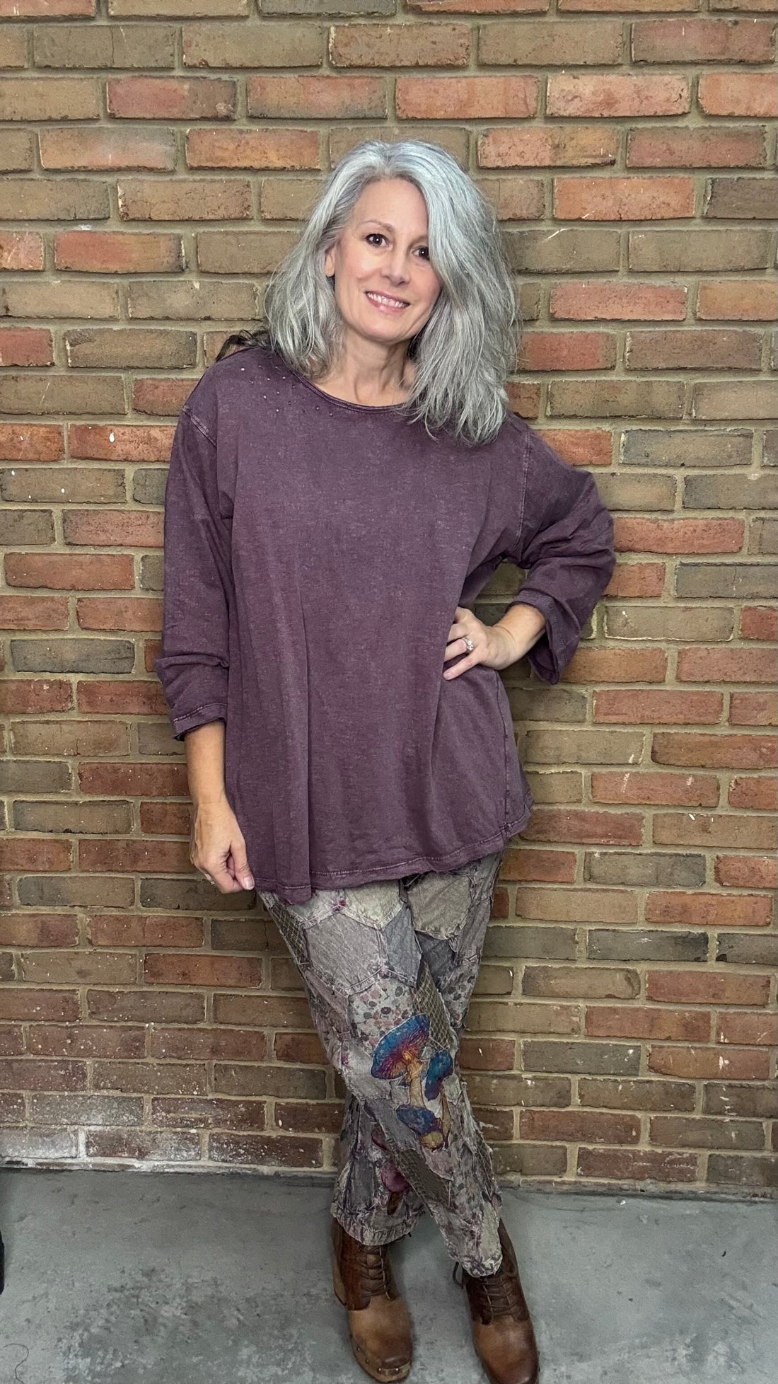 Person wearing a purple sweater and patterned pants standing against a brick wall.