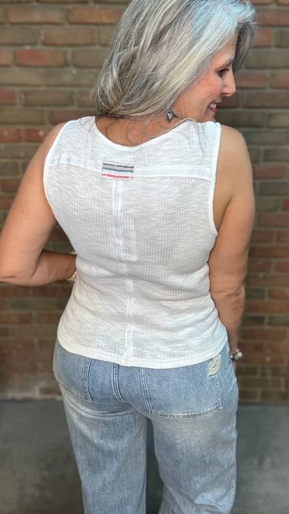 A woman with gray hair is shown from behind wearing a white sleeveless textured knit top and light blue jeans, standing against a brick wall.