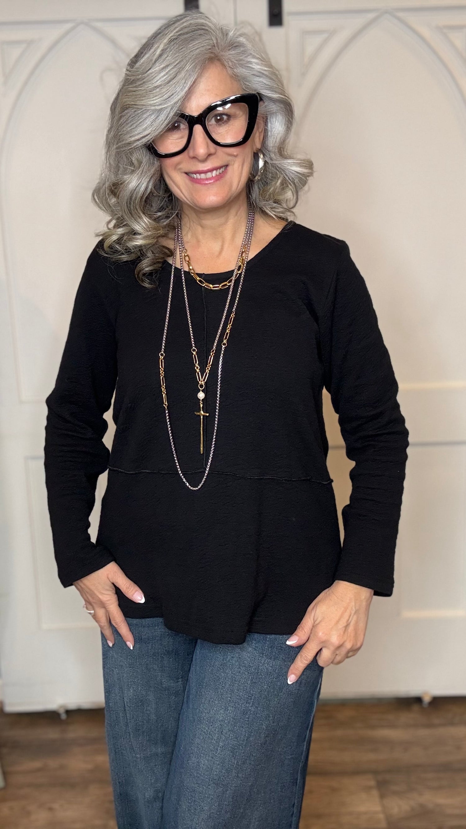 Woman wearing The Black Wanda Round Neck Top layered with the Silver Snake and Gold Paperclip Chain Necklace, the Silver Snake and Gold Paperclip Chain Choker Necklace and the Gold Cross w/Pearl Necklace  paired with  Judy Blue Palazzo jeans indoors against a white paneled wall.