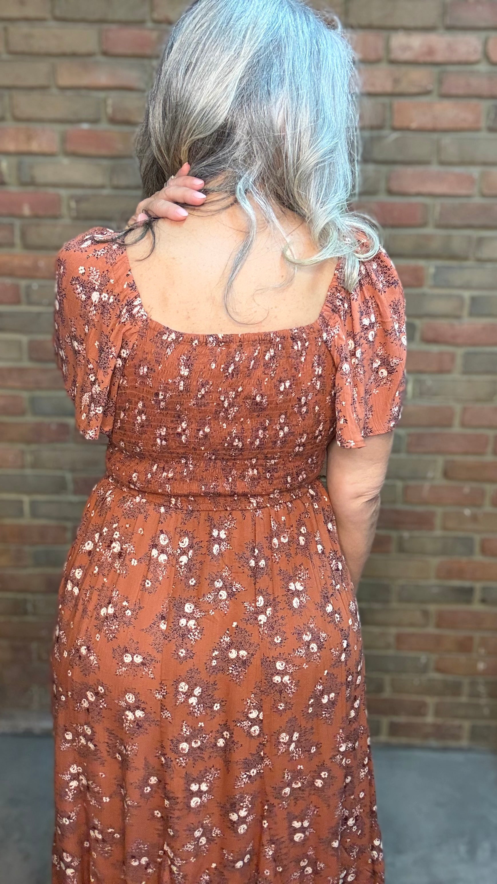 A woman with gray hair stands against a brick wall wearing a rust-brown bohemian-style midi dress with white floral prints, the "Short Sleeve Embroidered Midi Dress" and brown cowboy boots.