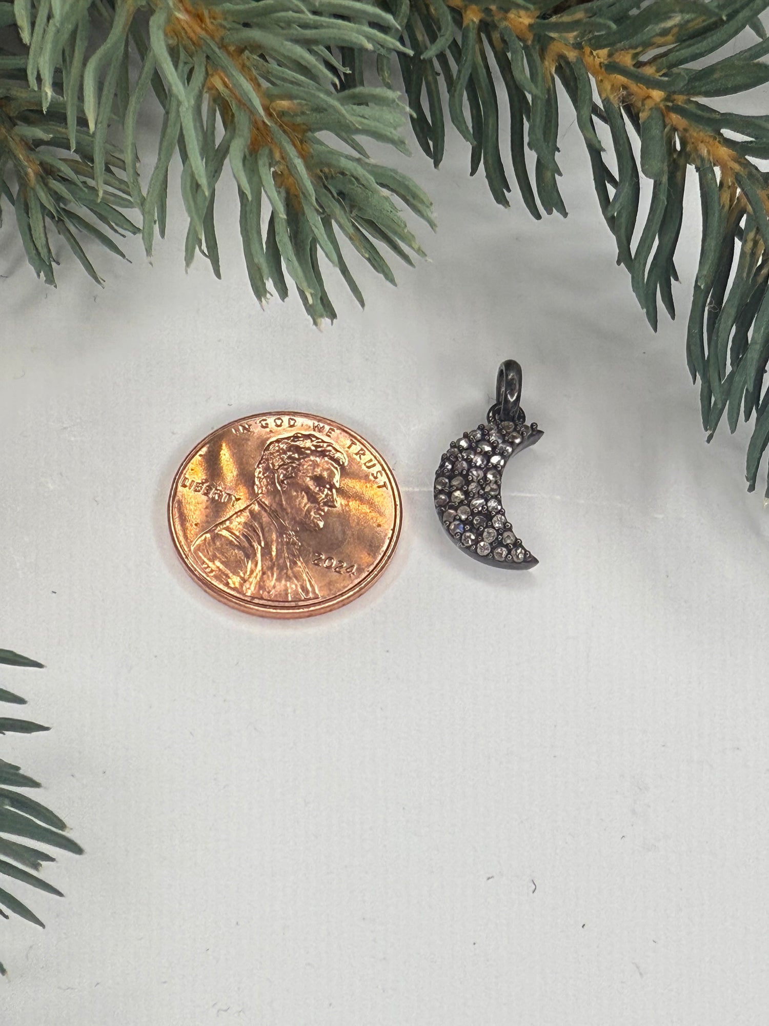 Small moon shaped pendant with pave diamonds next to a penny for scale on a white surface with greenery