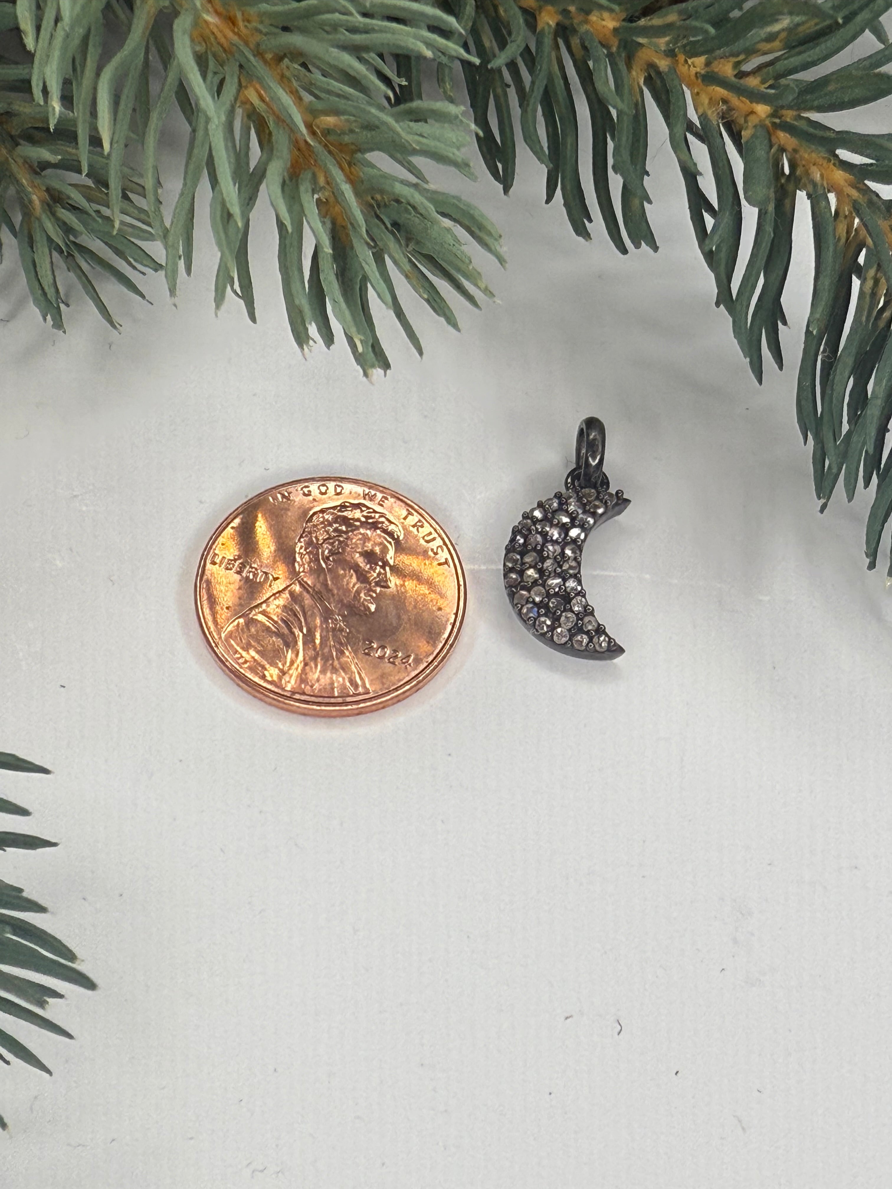 Small moon shaped pendant with pave diamonds next to a penny for scale on a white surface with greenery